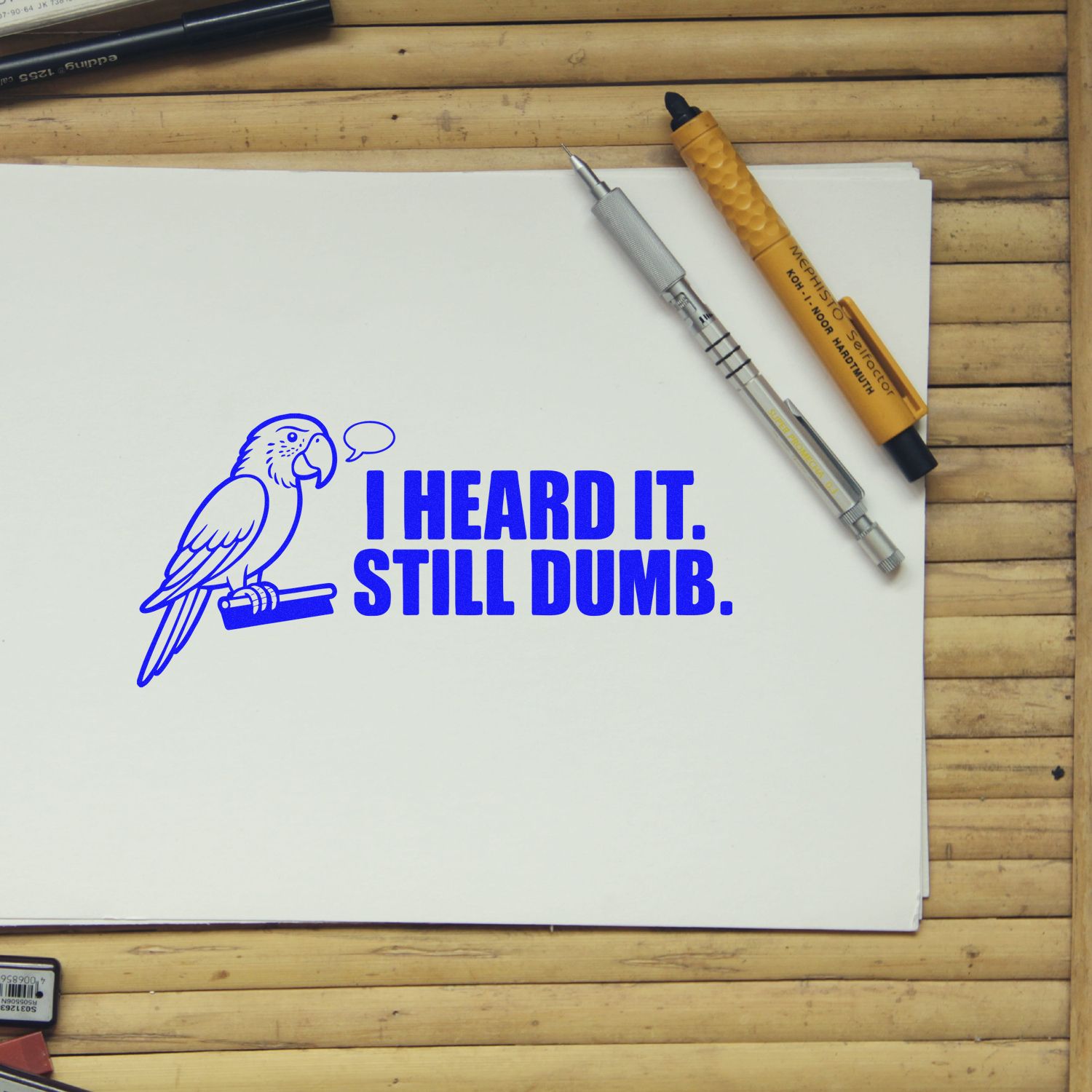 Blue parrot stamp reading I HEARD IT. STILL DUMB. on white paper, with silver and orange stamp and seal pens on a wood table.