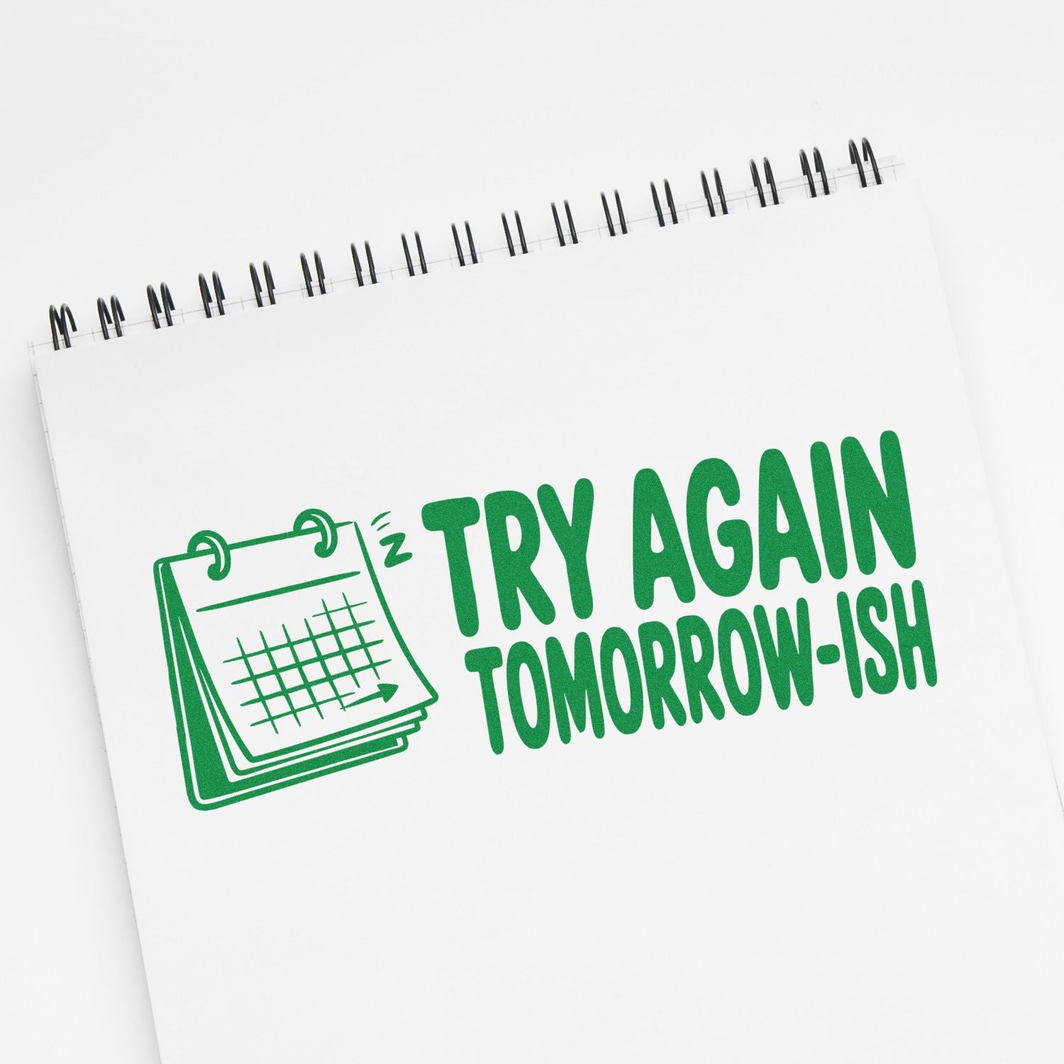 Green motivational stamp seal on white spiral notepad reading Try Again Tomorrow-ish with calendar icon.