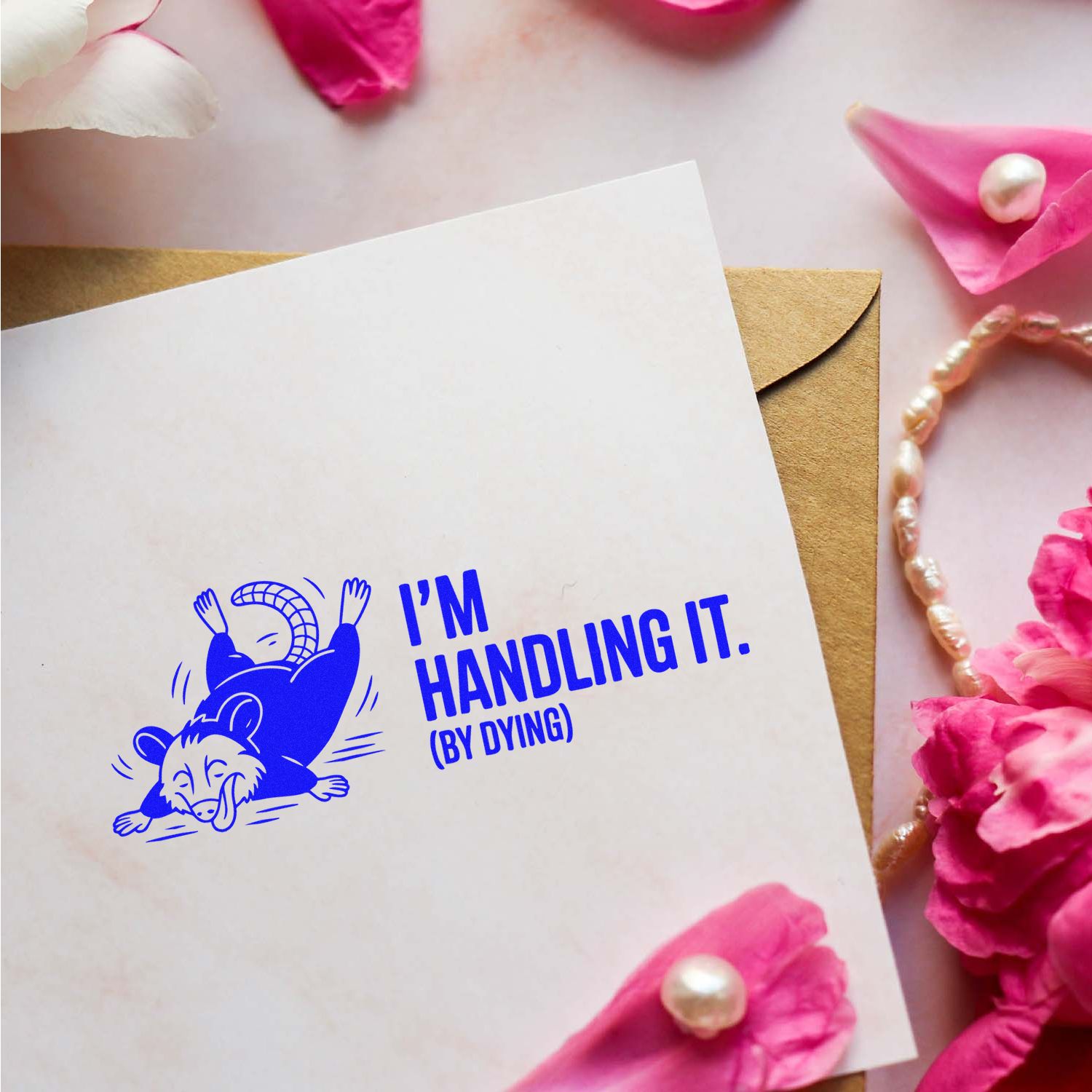 Blue opossum stamp reading Im handling it (by dying) on white card with kraft envelope, pink petals and pearls.