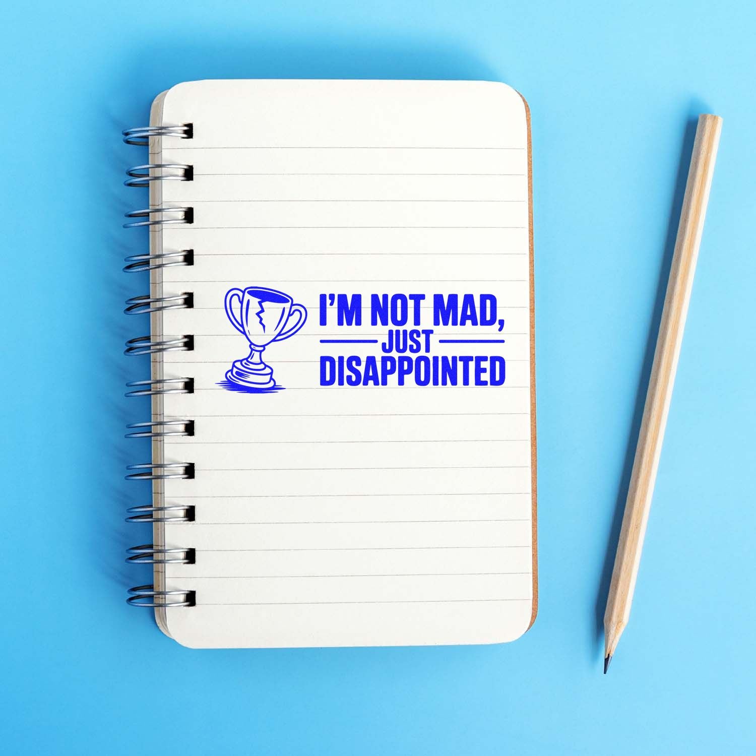 Blue self-inking stamp and seal with trophy icon and Im not mad, just disappointed on lined notebook, beside pencil.