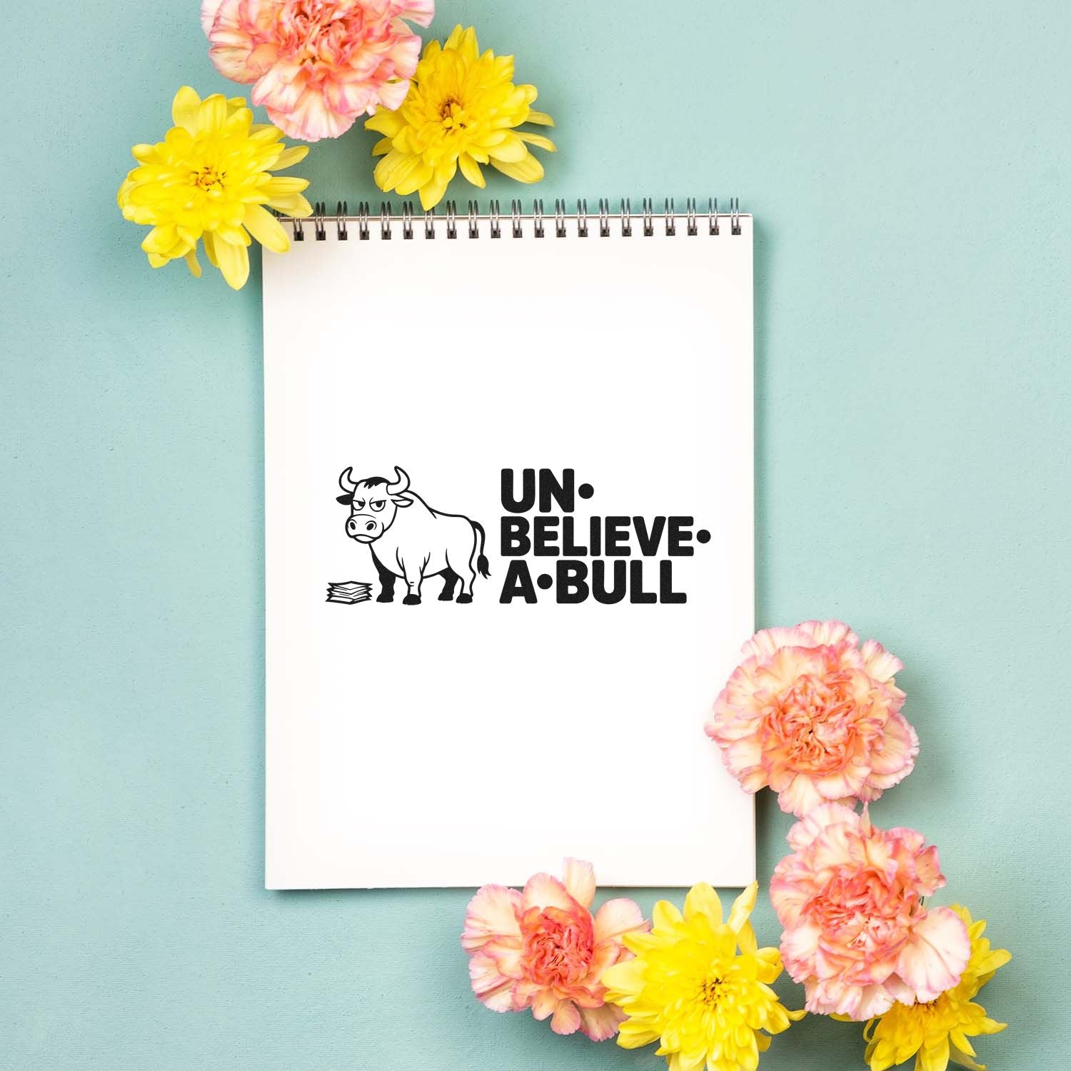 Black bull stamp and seal design reading UN-BELIEVE-A-BULL on a white spiral notebook, surrounded by yellow and pink flowers.