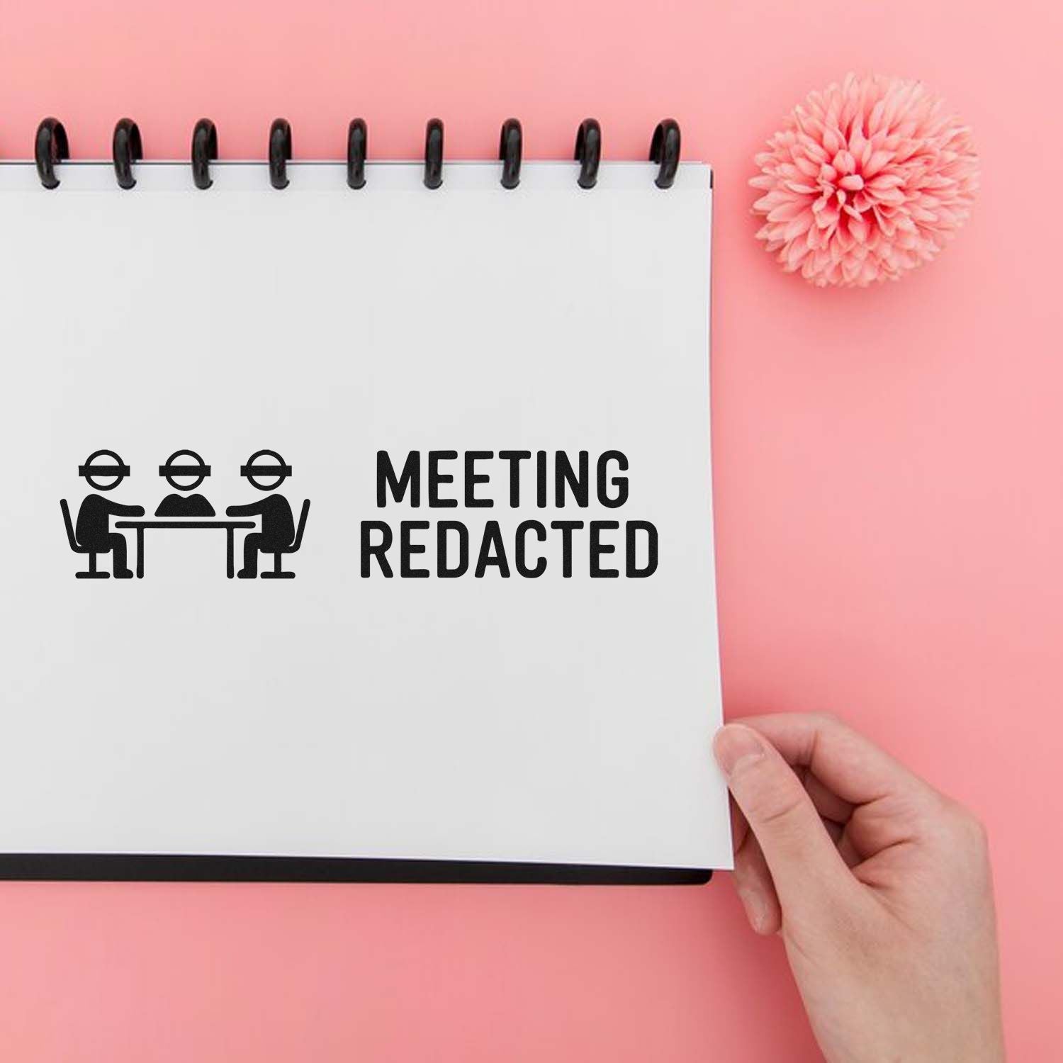 Black meeting redacted stamp on white spiral notebook, with hand, pink flower, and pastel pink background.