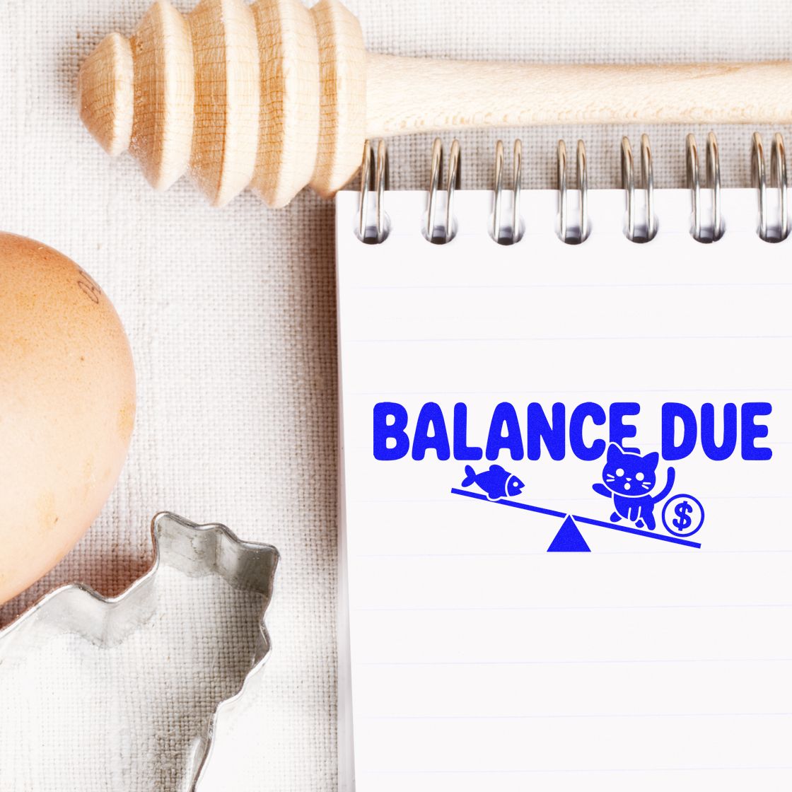 Blue BALANCE DUE stamp and seal on white notebook, with cat balance icon, dollar sign, and beige wooden stamp tools.