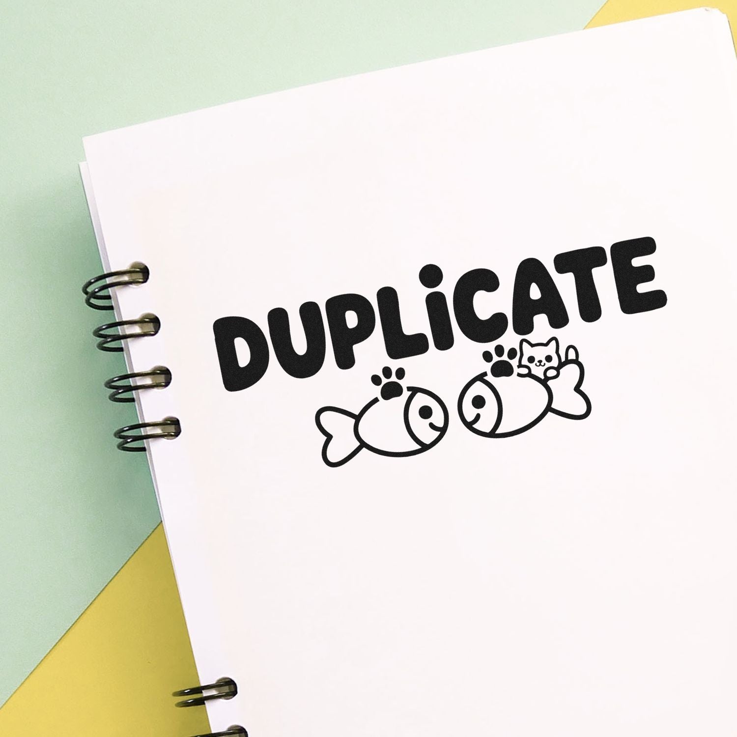 Black DUPLICATE stamp seal with fish and cat icon on a white spiral notebook, pastel green and yellow background.