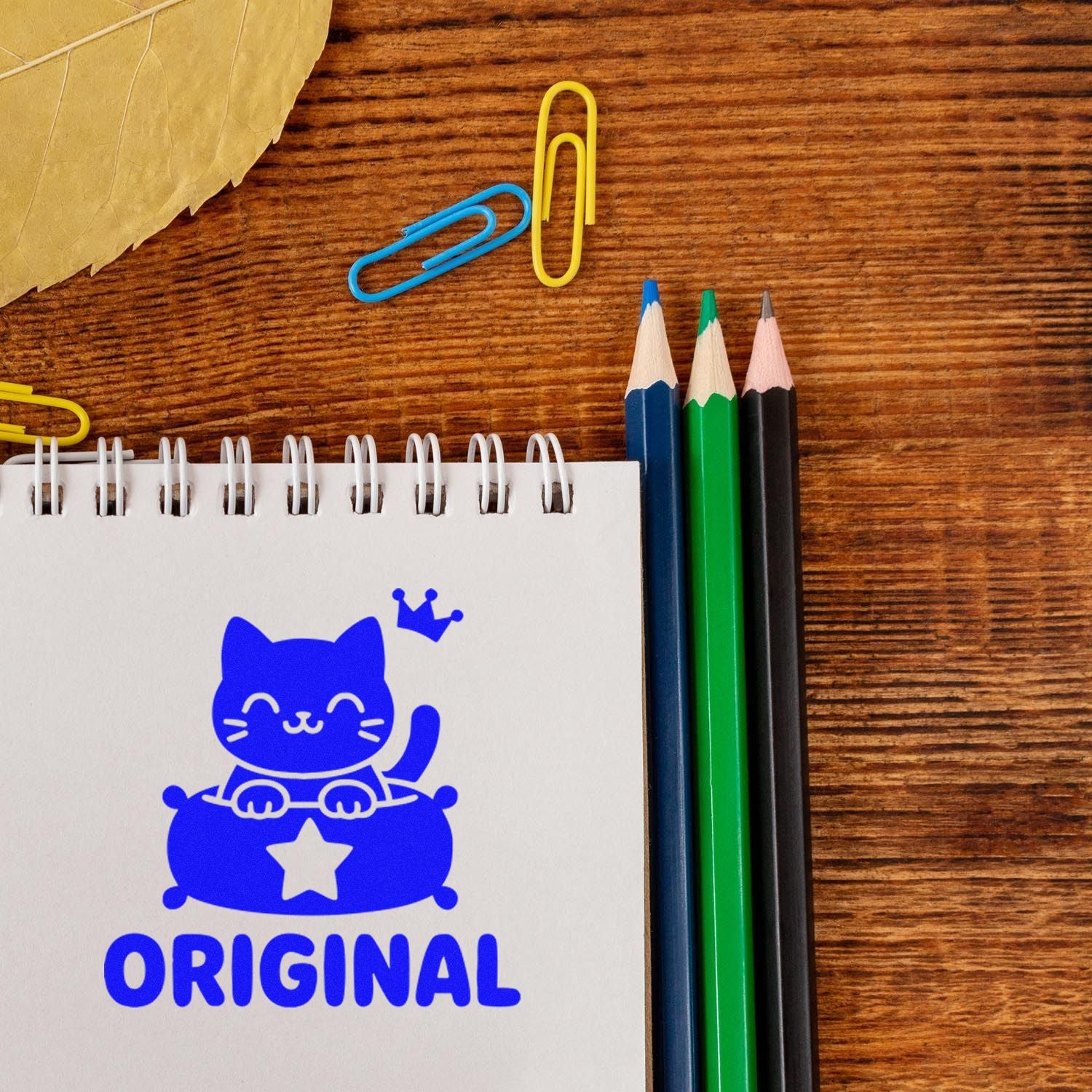 Blue cat ORIGINAL stamp and seal on a spiral notebook, with colored pencils, paper clips, and a wood desk.
