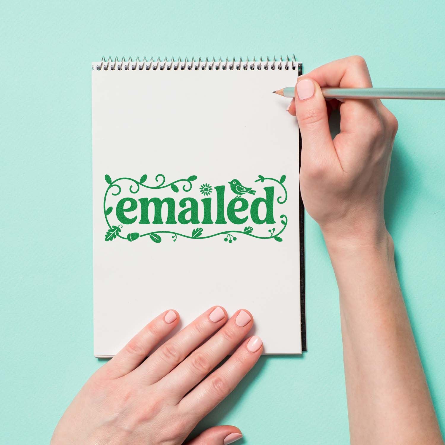 Green decorative emailed stamp seal on a white spiral notebook, with hands holding a pencil.