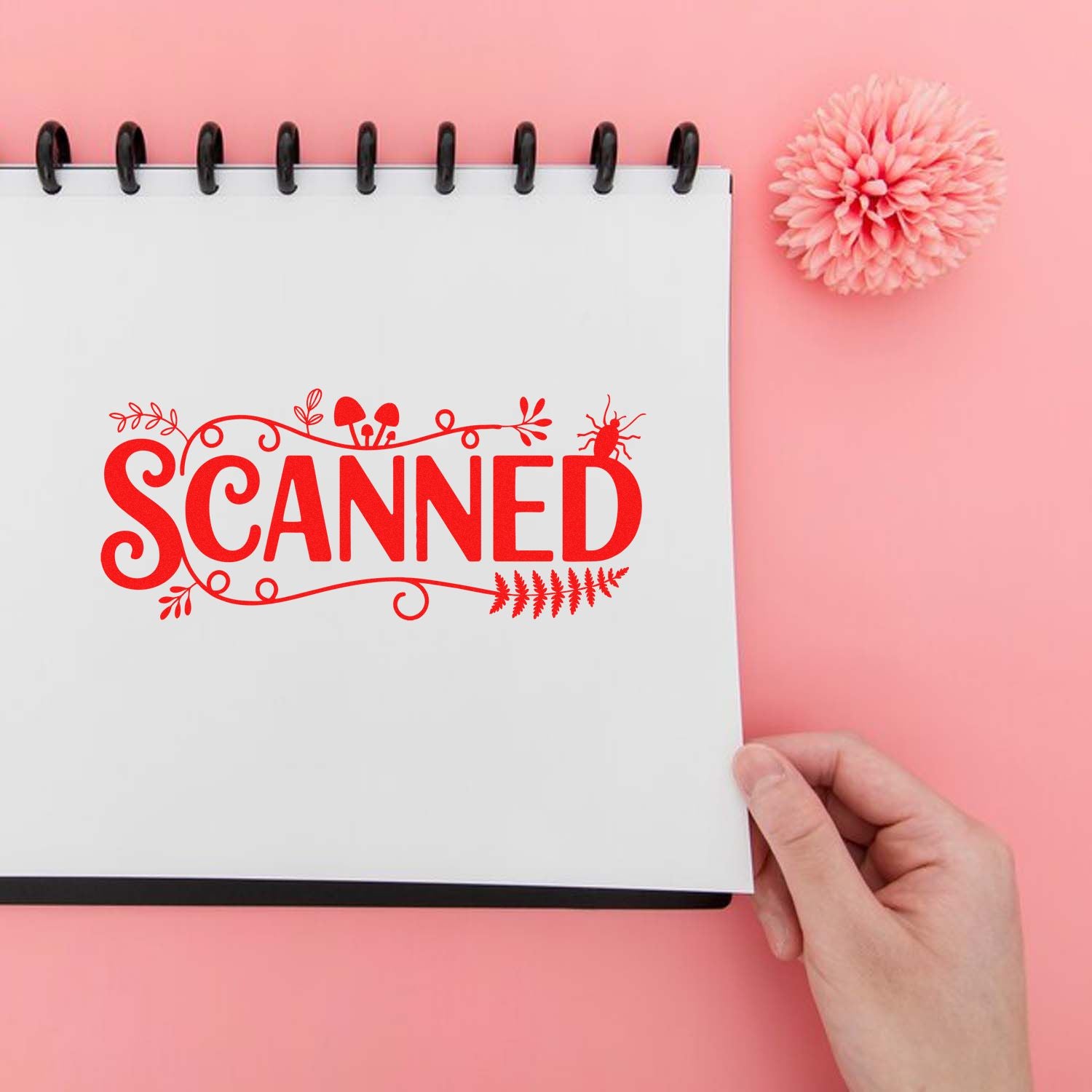 Red decorative SCANNED stamp seal on a white spiral notebook, with a pink background, hand, and pink flower.
