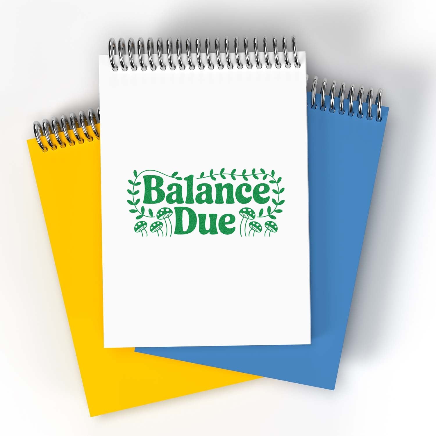 White spiral notepad with green Balance Due seal stamp, leafy mushrooms, over yellow and blue notebooks.