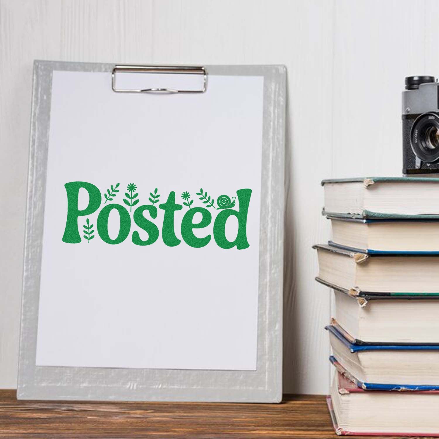 Green Posted stamp design on clipboard, with leaf and snail accents beside stacked books and a camera.