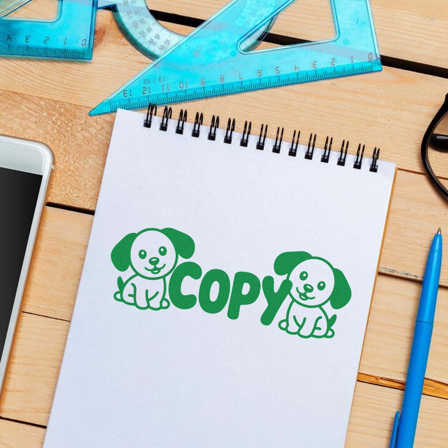Green dog-themed Copy stamp seal on a white spiral notebook, with blue rulers and pen on a wooden desk.