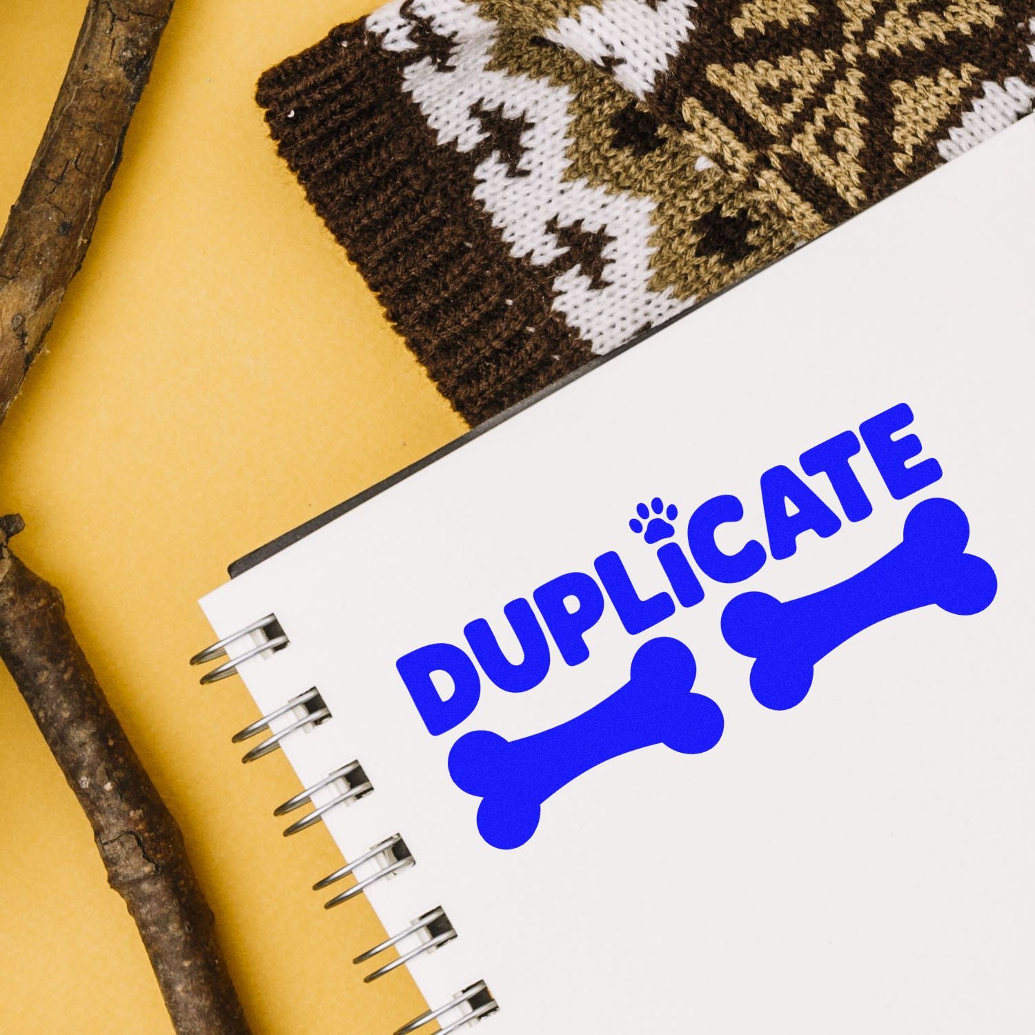 Blue duplicate stamp and seal with bone icons on a white spiral notebook, set on mustard yellow with knit and wood textures.