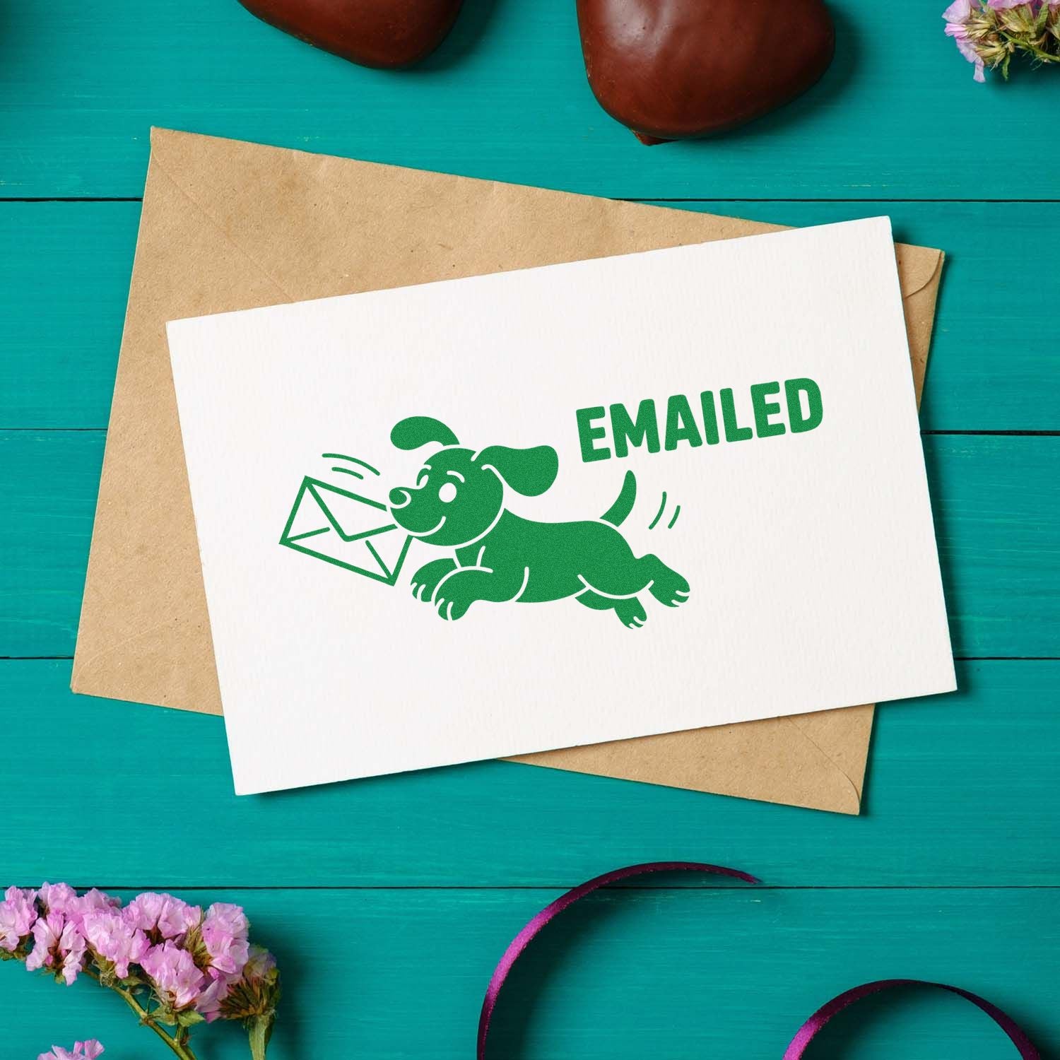 Green EMAILED stamp seal with dog carrying envelope on white card, layered over a kraft envelope on teal wood.
