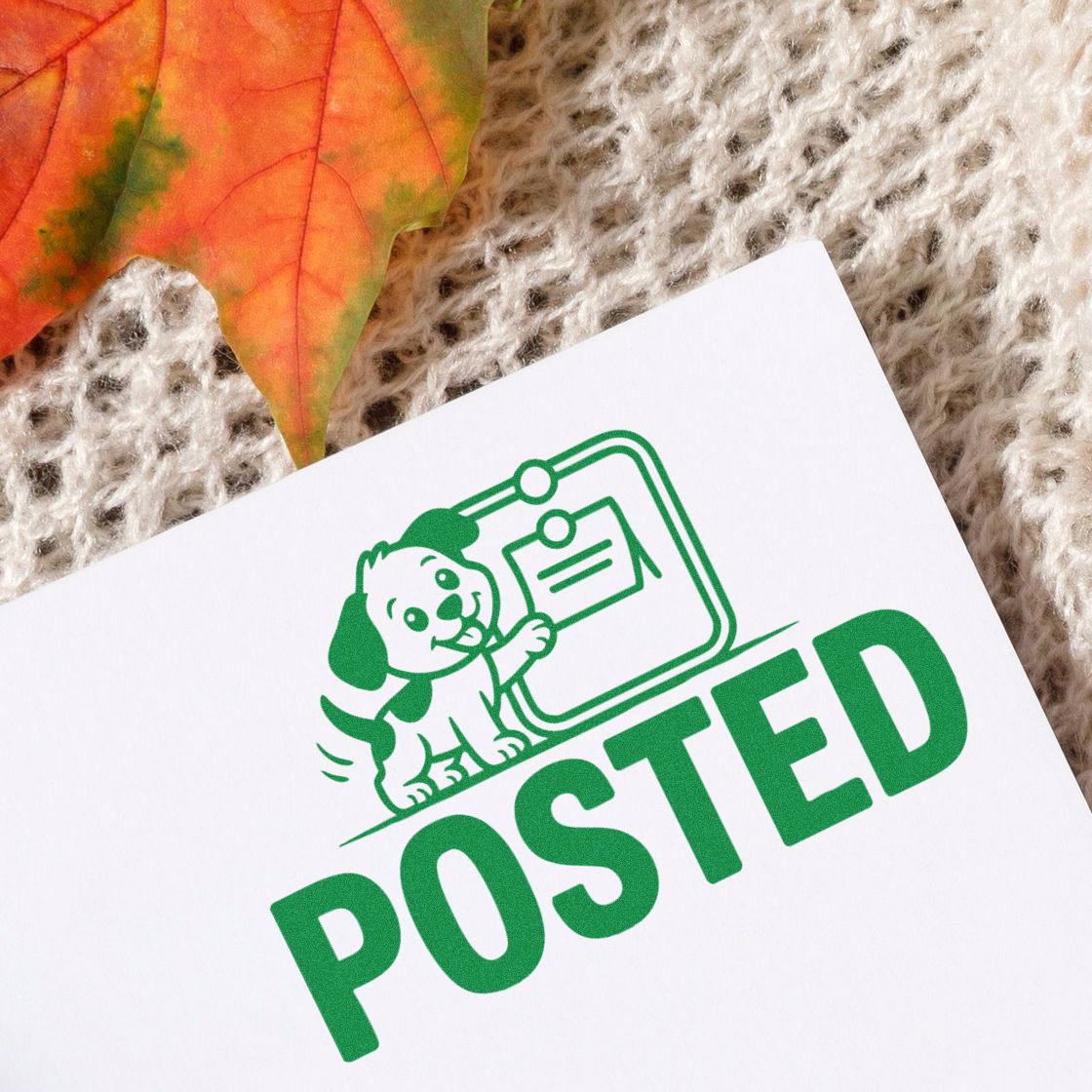 Green POSTED stamp seal with dog mascot on white paper, beside an orange autumn leaf on beige knit fabric.