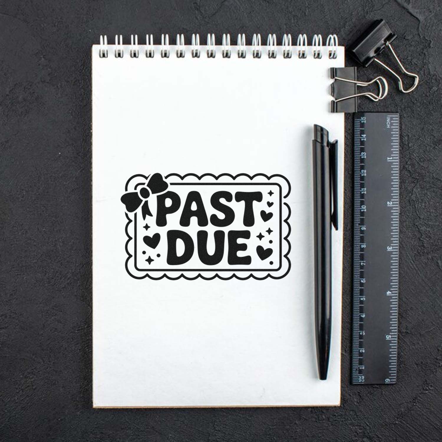 Black PAST DUE stamp seal with hearts and bow on white notepad, beside a black pen, ruler, and binder clips.