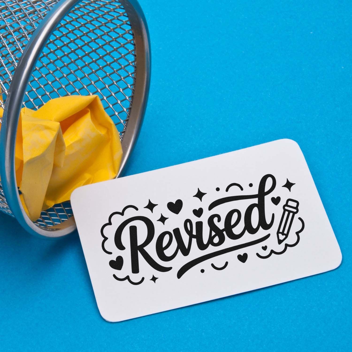 White revised stamp and seal card with black script, hearts, and pencil on blue background beside a mesh basket with yellow tissue.