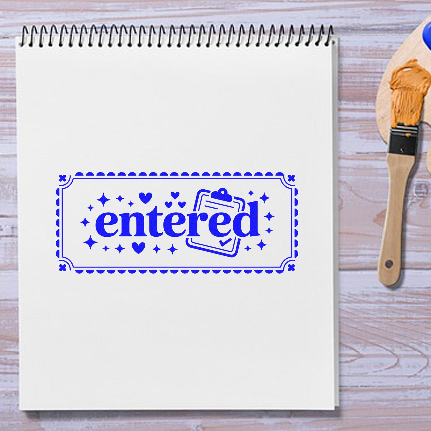 White spiral notebook with bright blue entered stamp seal, hearts and clipboard icon, beside a wooden rubber stamp brush.