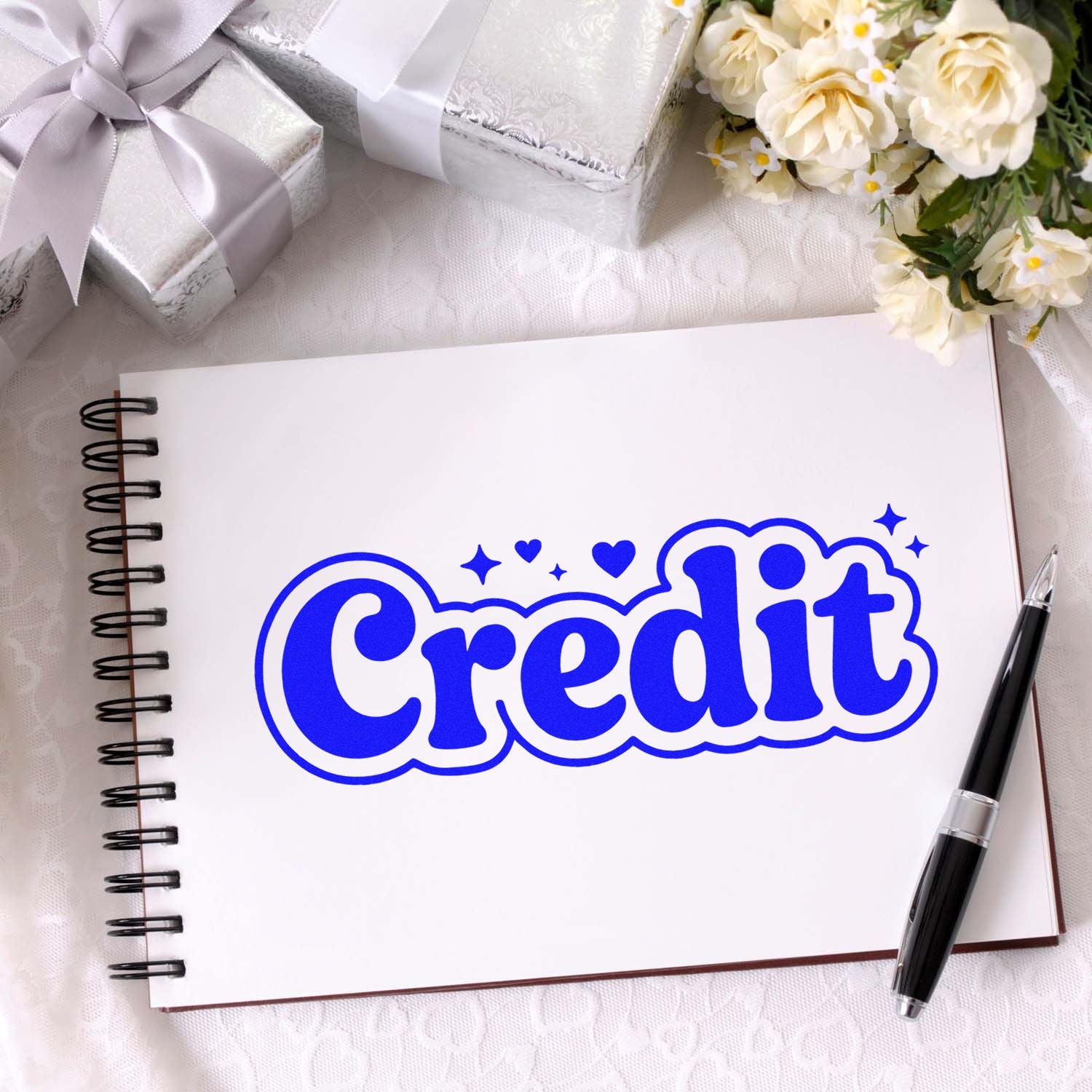 Blue Credit stamp and seal on white spiral notebook, surrounded by silver gift boxes, white flowers, and a black pen