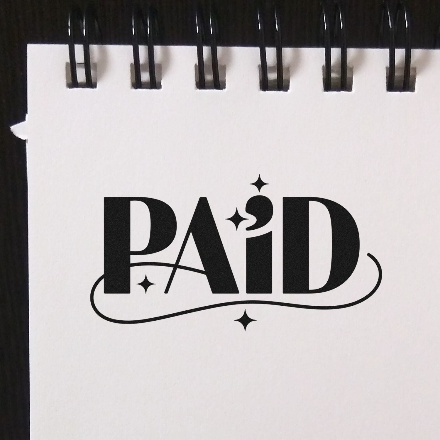 Black PAID stamp and seal design on a white spiral-bound page with sparkles and a curved underline.