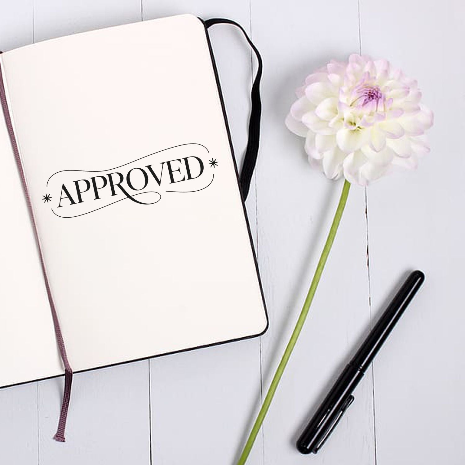 Notebook with black APPROVED stamp and seal, white flower, and black pen on a white wood background.