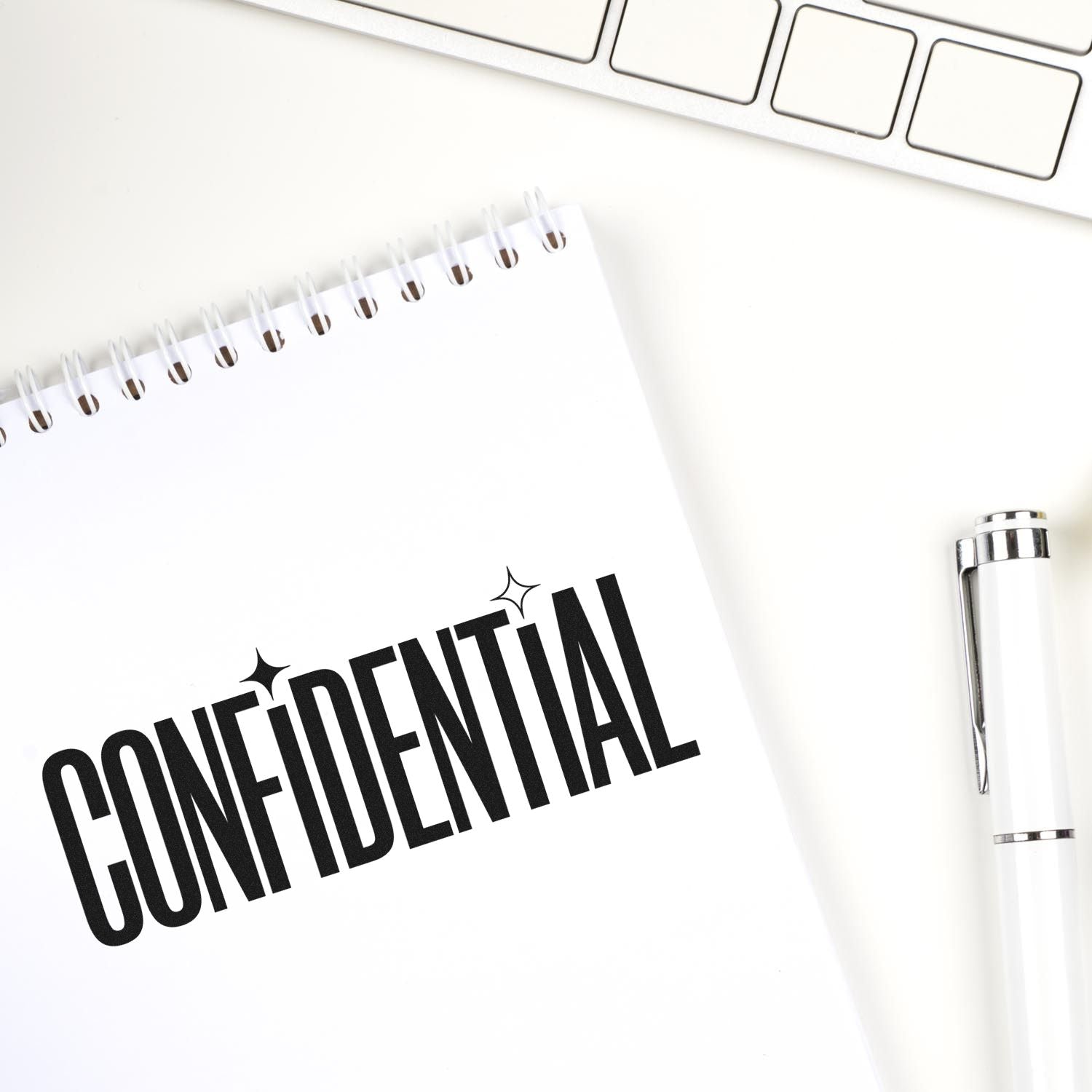 White notebook with bold black CONFIDENTIAL stamp seal, silver pen, and keyboard on a clean desk.