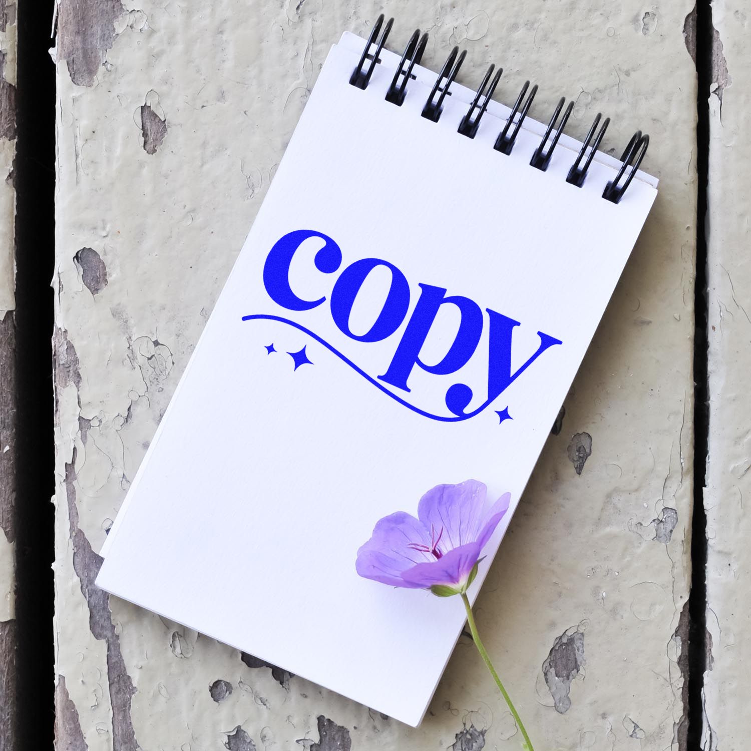 Blue "copy" stamp and seal on a white spiral notebook, with a purple flower on weathered gray wood.