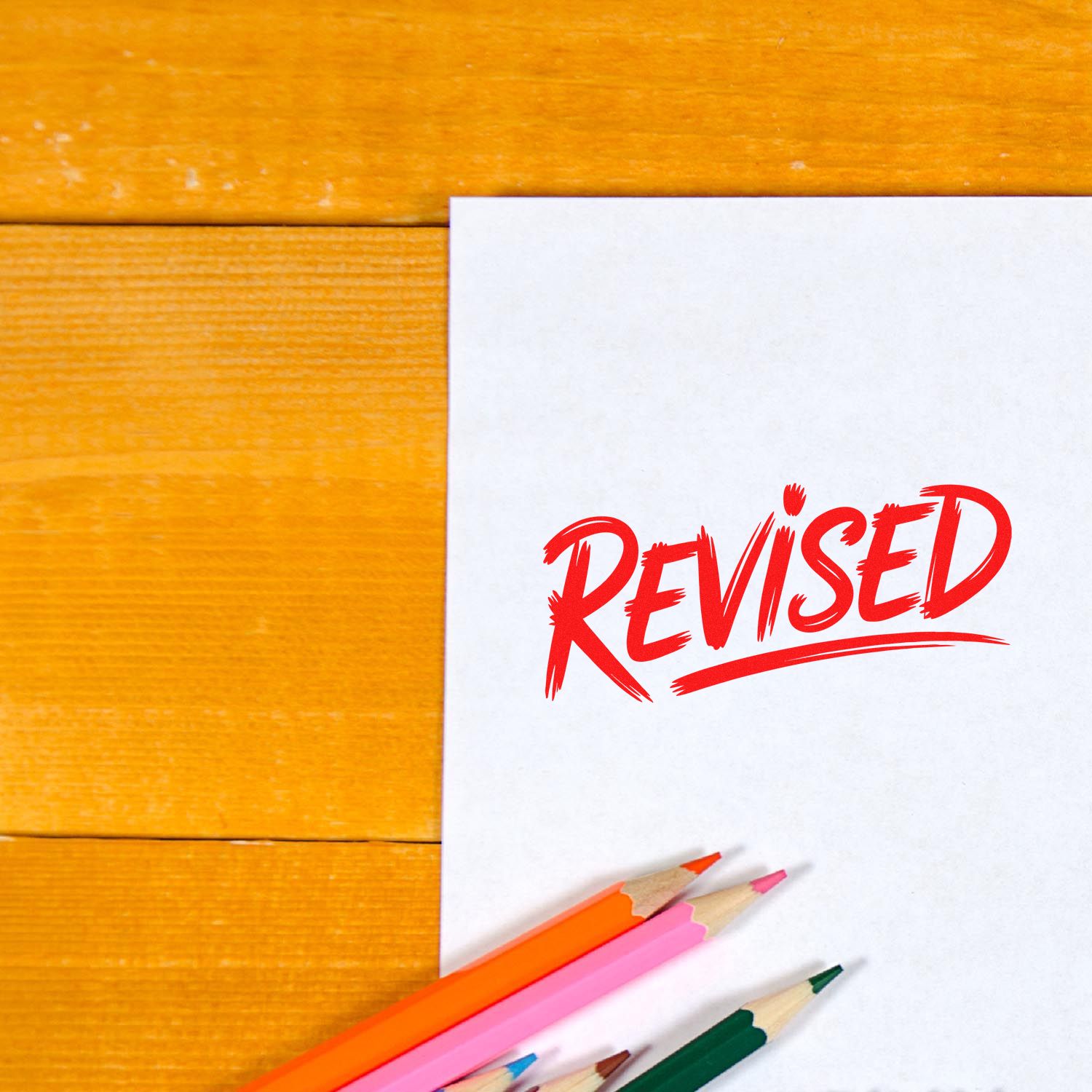 White paper with red REVISED stamp and colored pencils on a wooden desk, showcasing seal and stamp products.