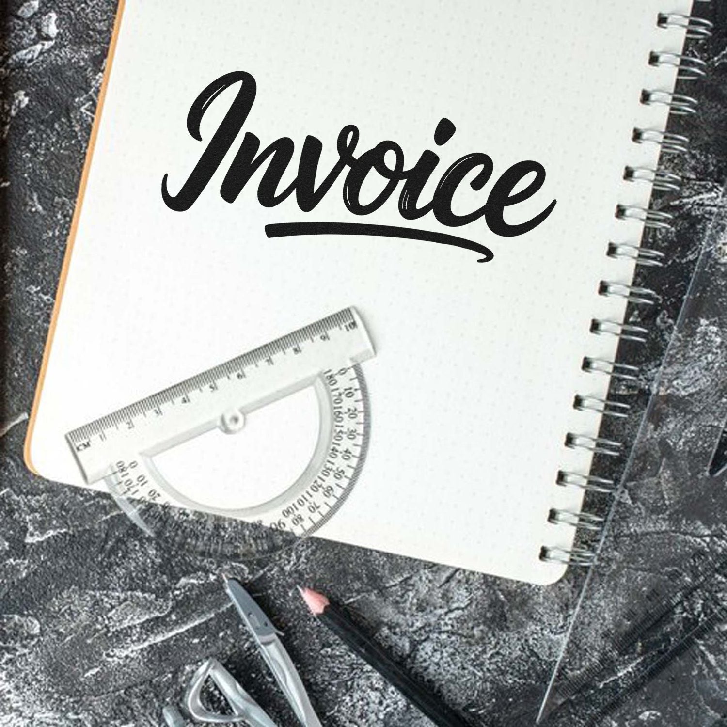Black invoice stamp and seal on a white spiral notebook, with clear protractor and drafting tools on a dark textured background.