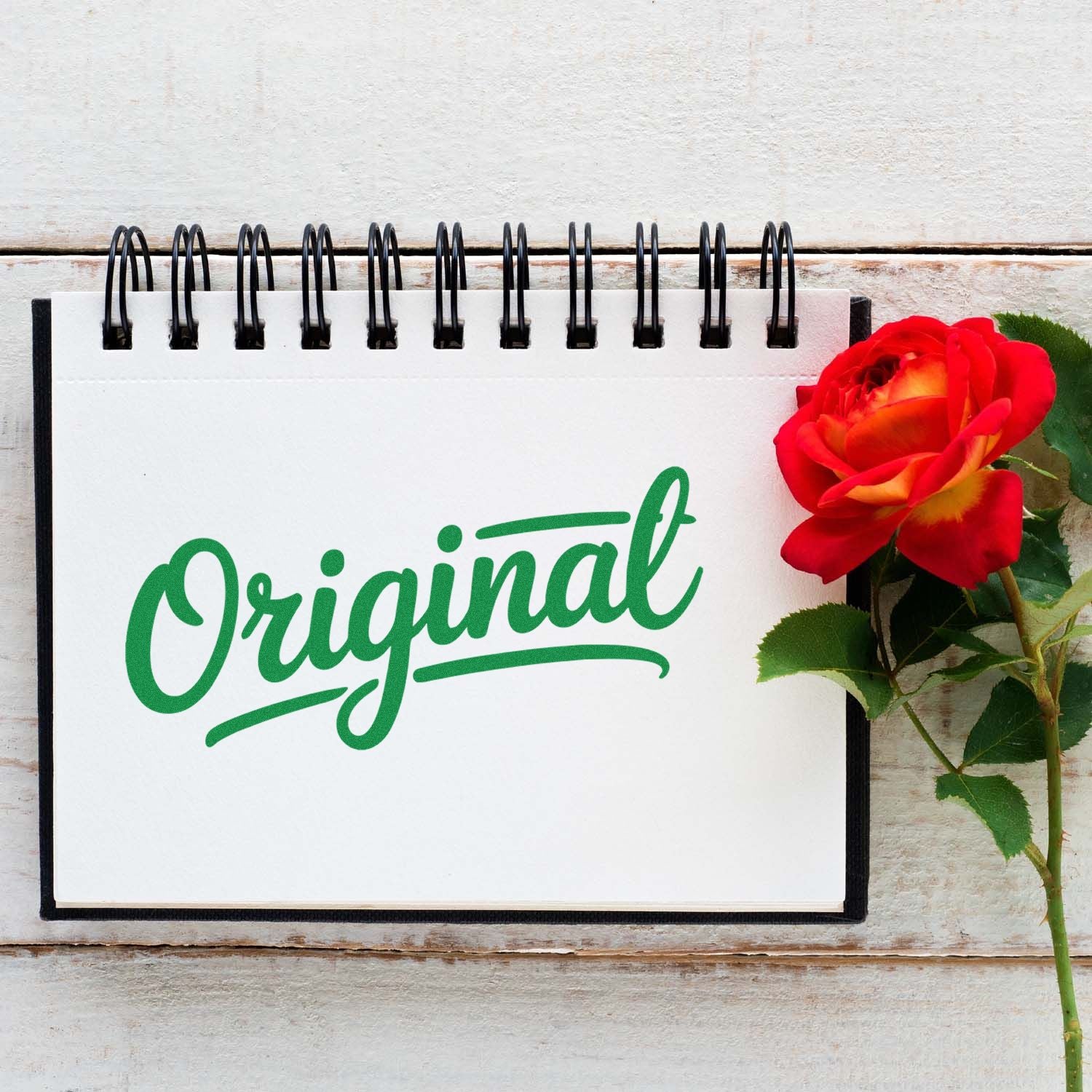 Green Original stamp and seal design on a white spiral notebook beside a red rose.