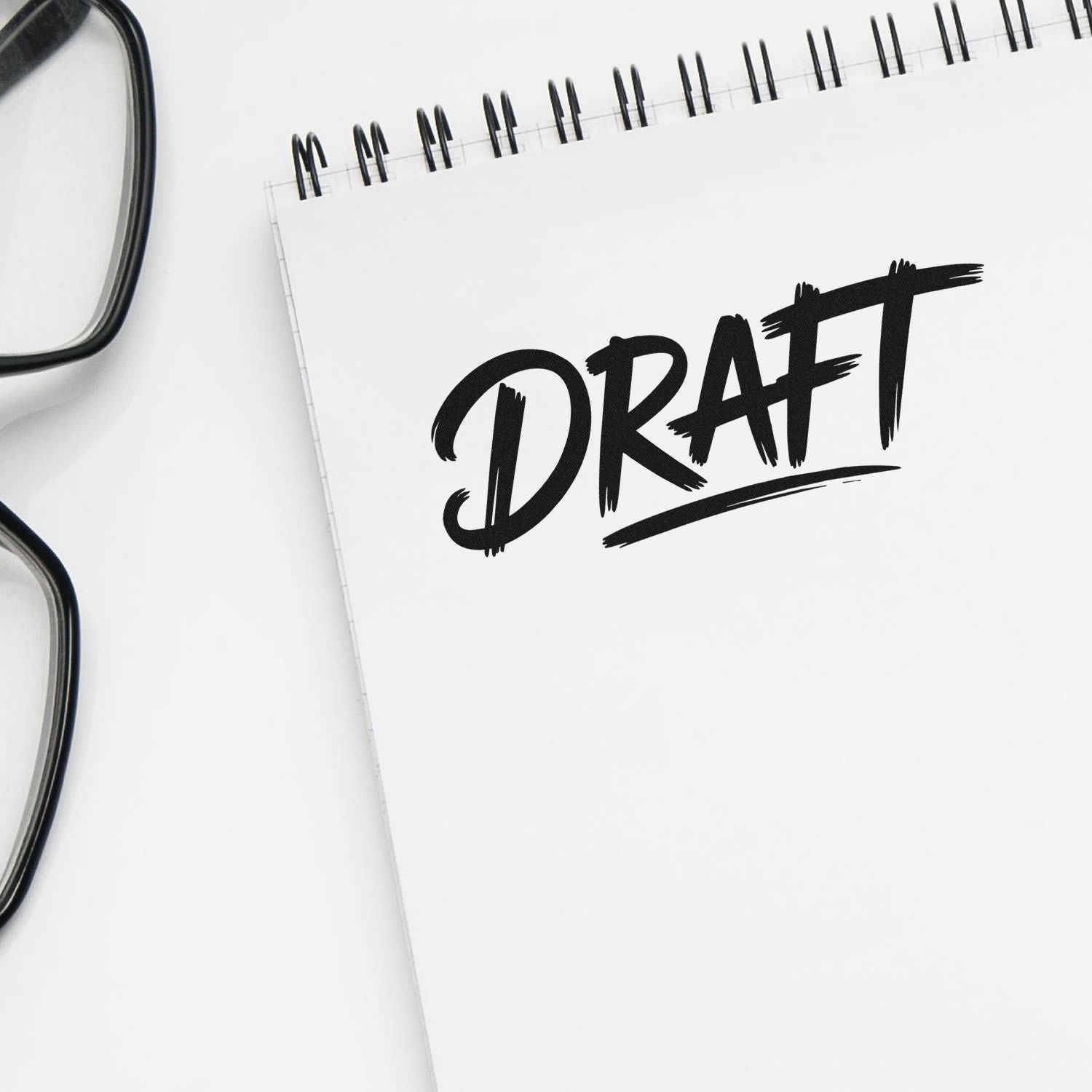 Black DRAFT stamp on white spiral notepad beside eyeglasses, showing a custom seal for document approval.