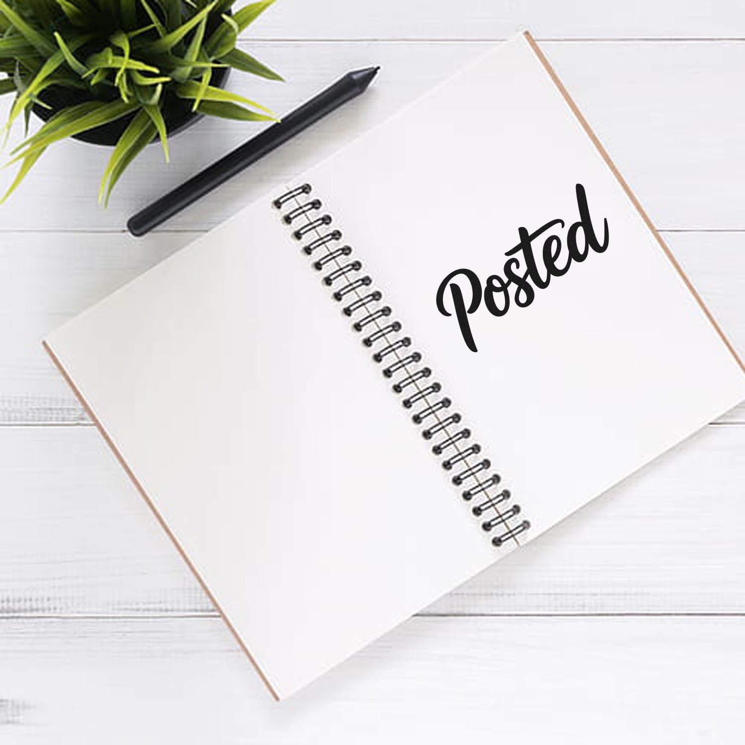 White spiral notebook with black Posted stamp imprint, black pen, and green plant on a white wood desk.