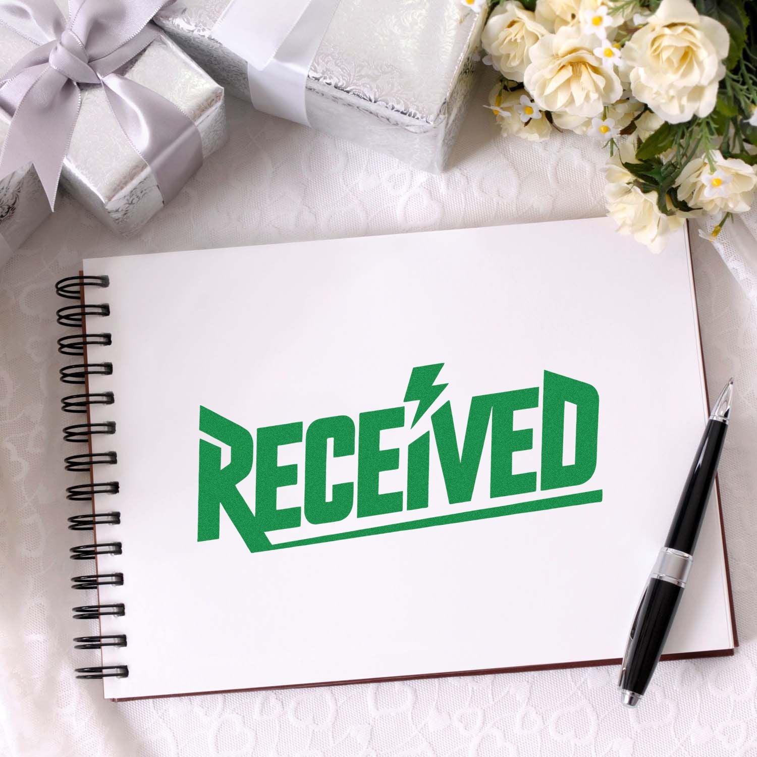 Green RECEIVED stamp on white spiral notebook, with silver gift boxes, white flowers, and a black pen.