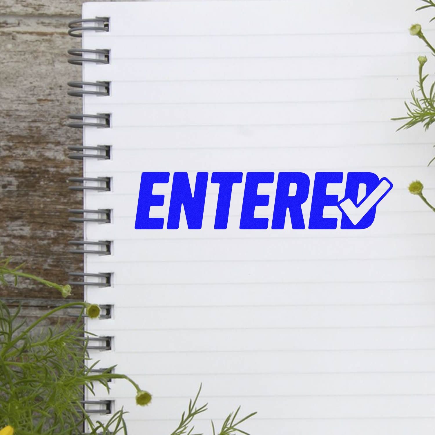 Blue ENTERED rubber stamp with checkmark on white spiral notebook, framed by greenery.