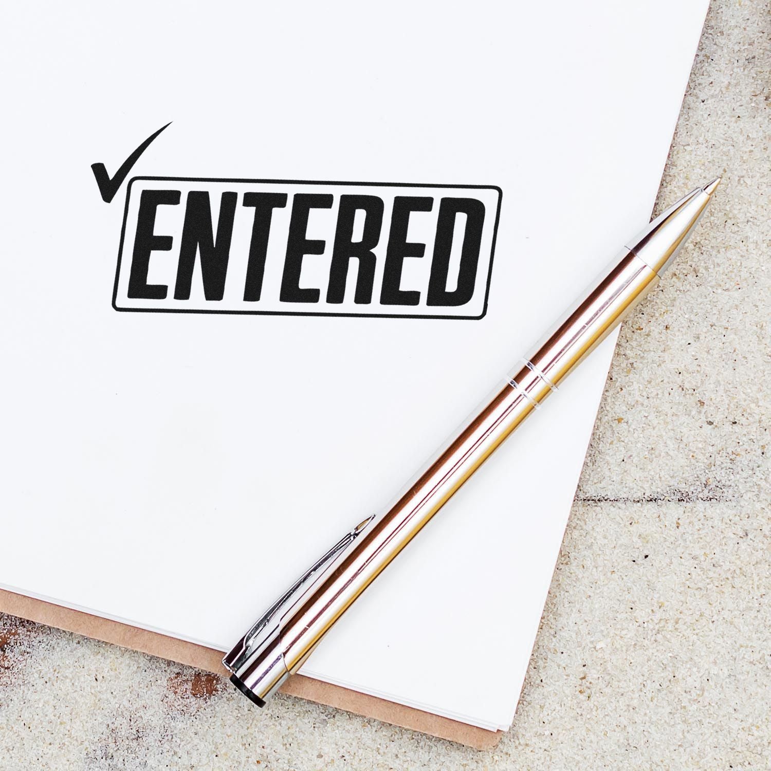 Black ENTERED rubber stamp seal with checkmark on white paper, beside a gold-and-rose pen.