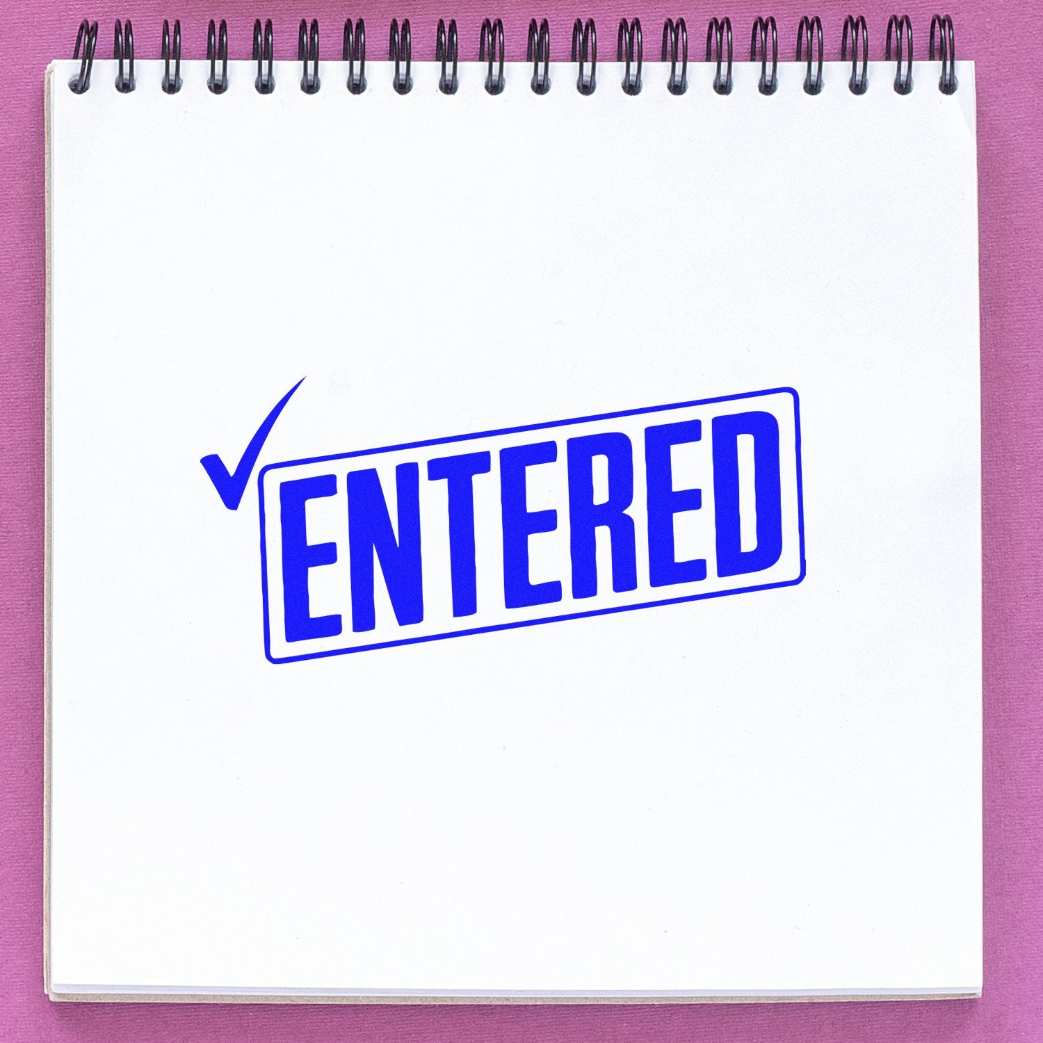 Blue ENTERED rubber stamp with checkmark on white spiral notepad over pink background.