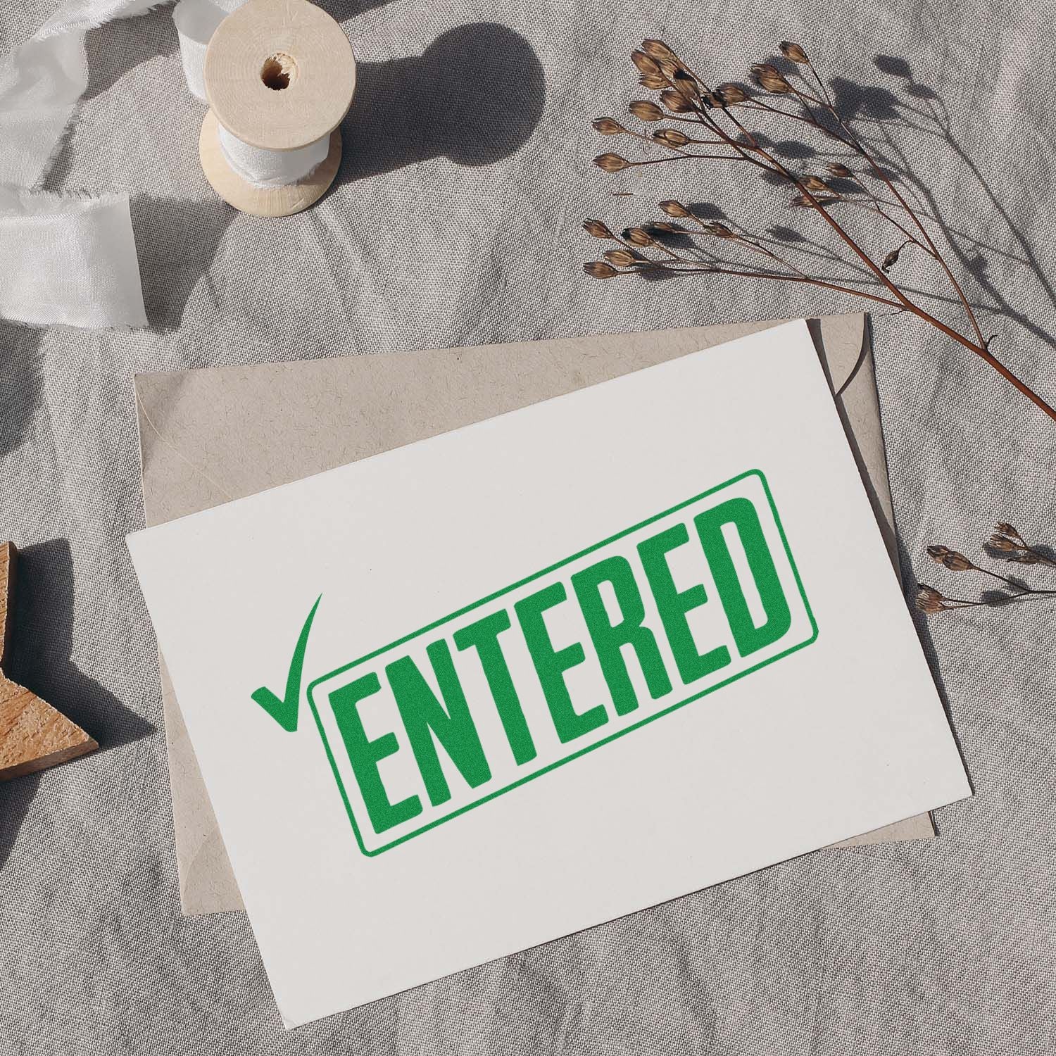 Green ENTERED stamp seal on white card atop beige envelope, with ribbon spool and dried stems.