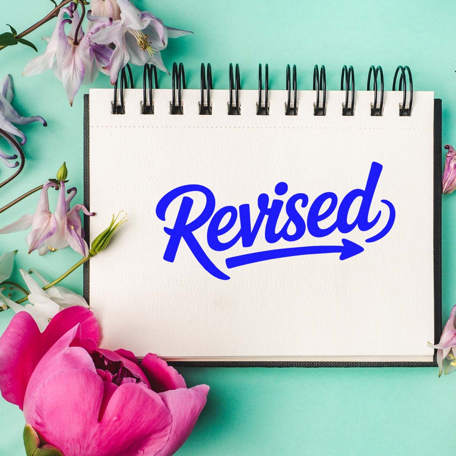 Blue "Revised" stamp and seal mark on white spiral notebook, with pink and purple flowers on mint green background.