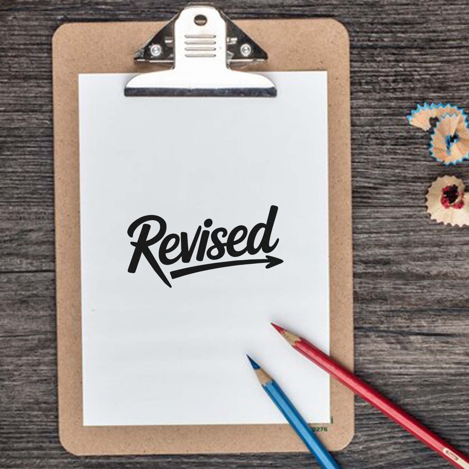Black "Revised" self-inking stamp on white clipboard, with red and blue pencils on a wood desk.