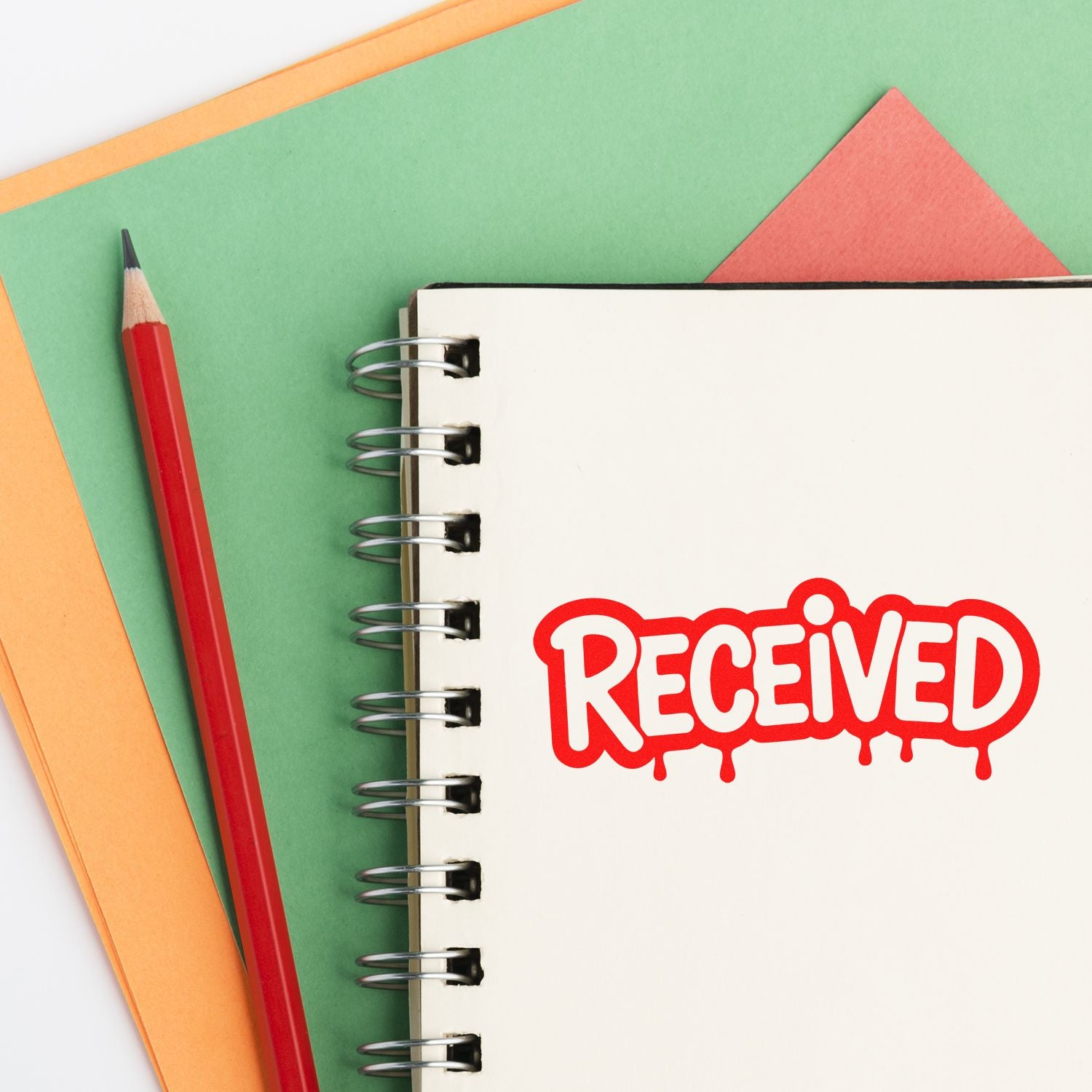 Red RECEIVED stamp seal on white spiral notebook beside a red pencil, with green and orange paper.