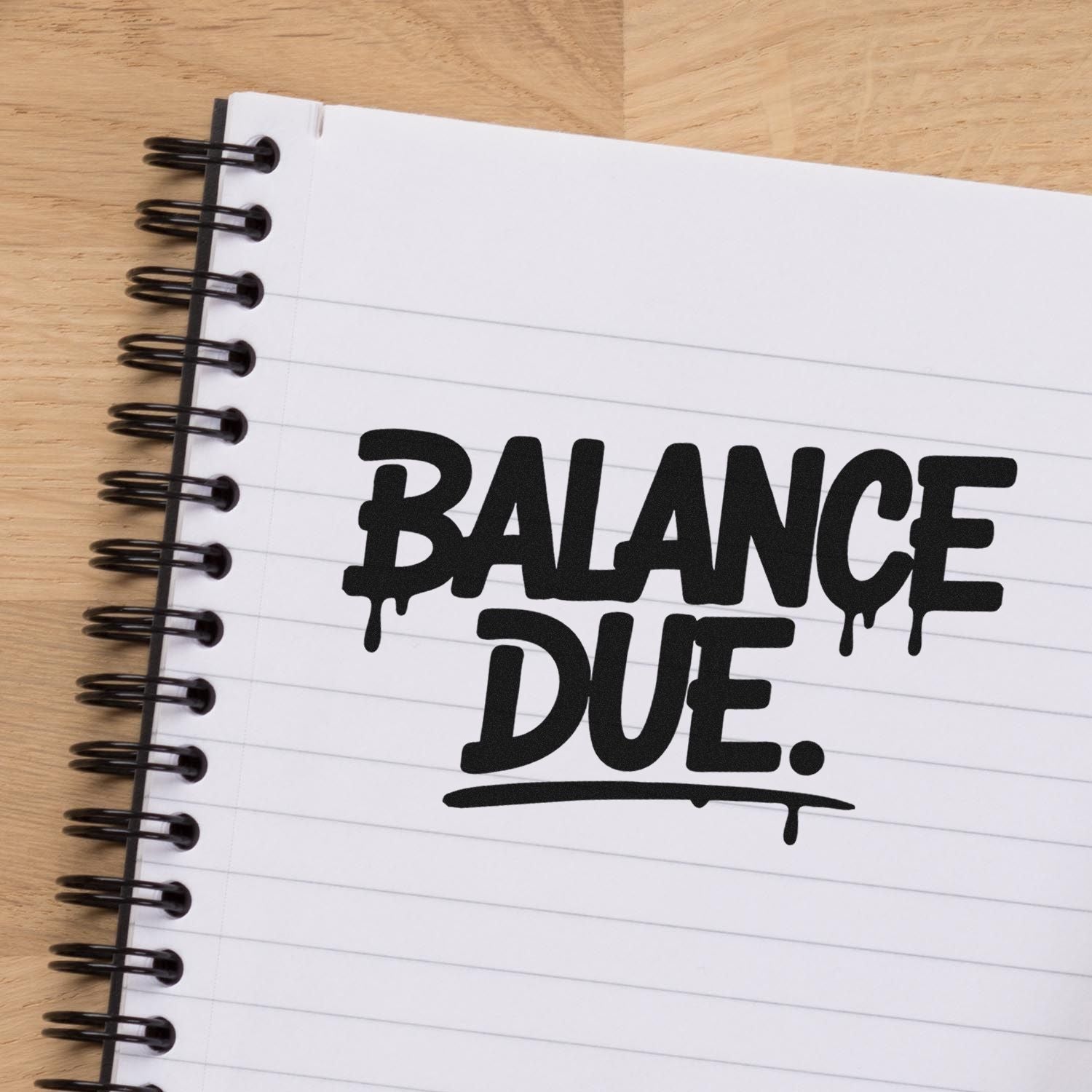Black BALANCE DUE stamp-style seal on white spiral notebook over wood desk.