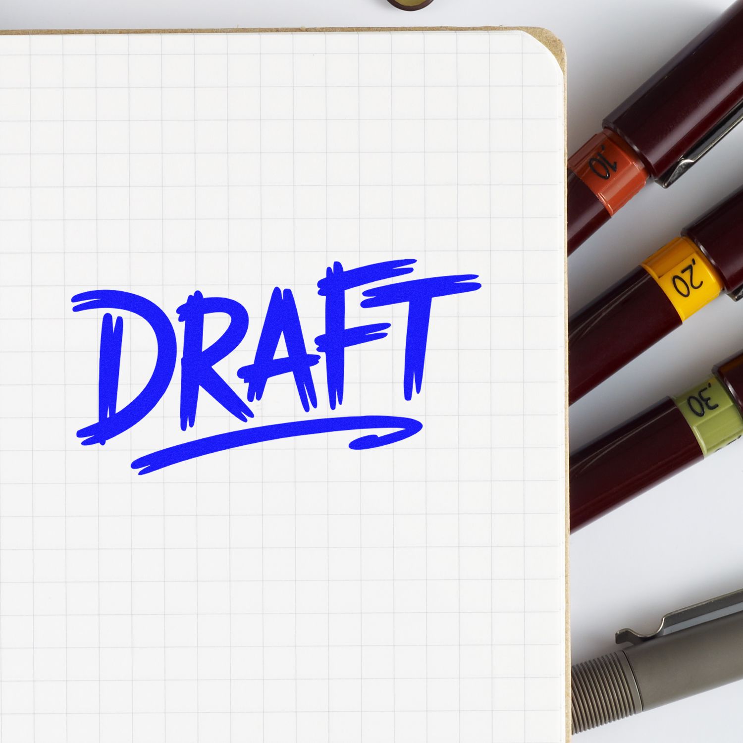 Blue DRAFT on graph paper notebook beside burgundy stamp and seal pens with orange, yellow, and green bands.