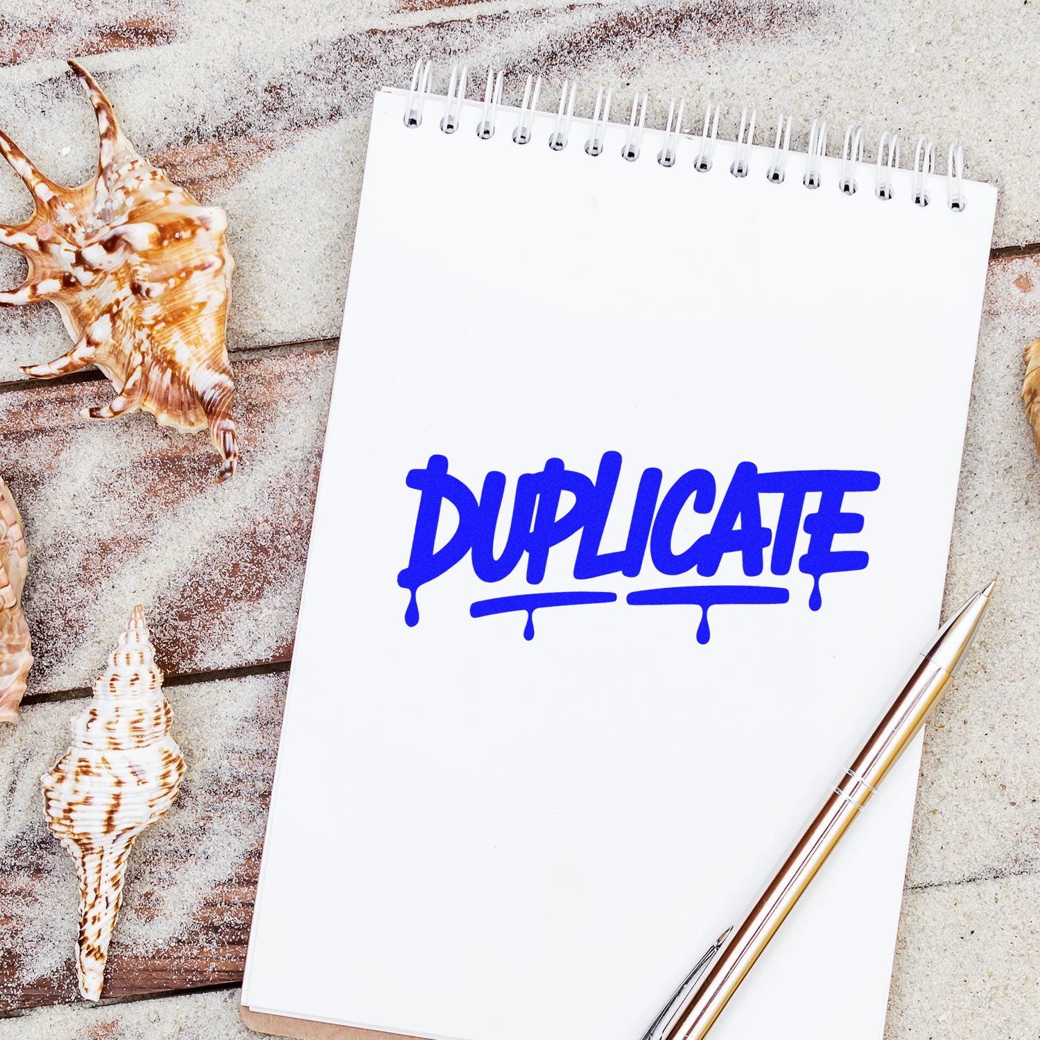 Blue DUPLICATE stamp on white spiral notebook, beside seashells and a gold pen on a sandy wood background.