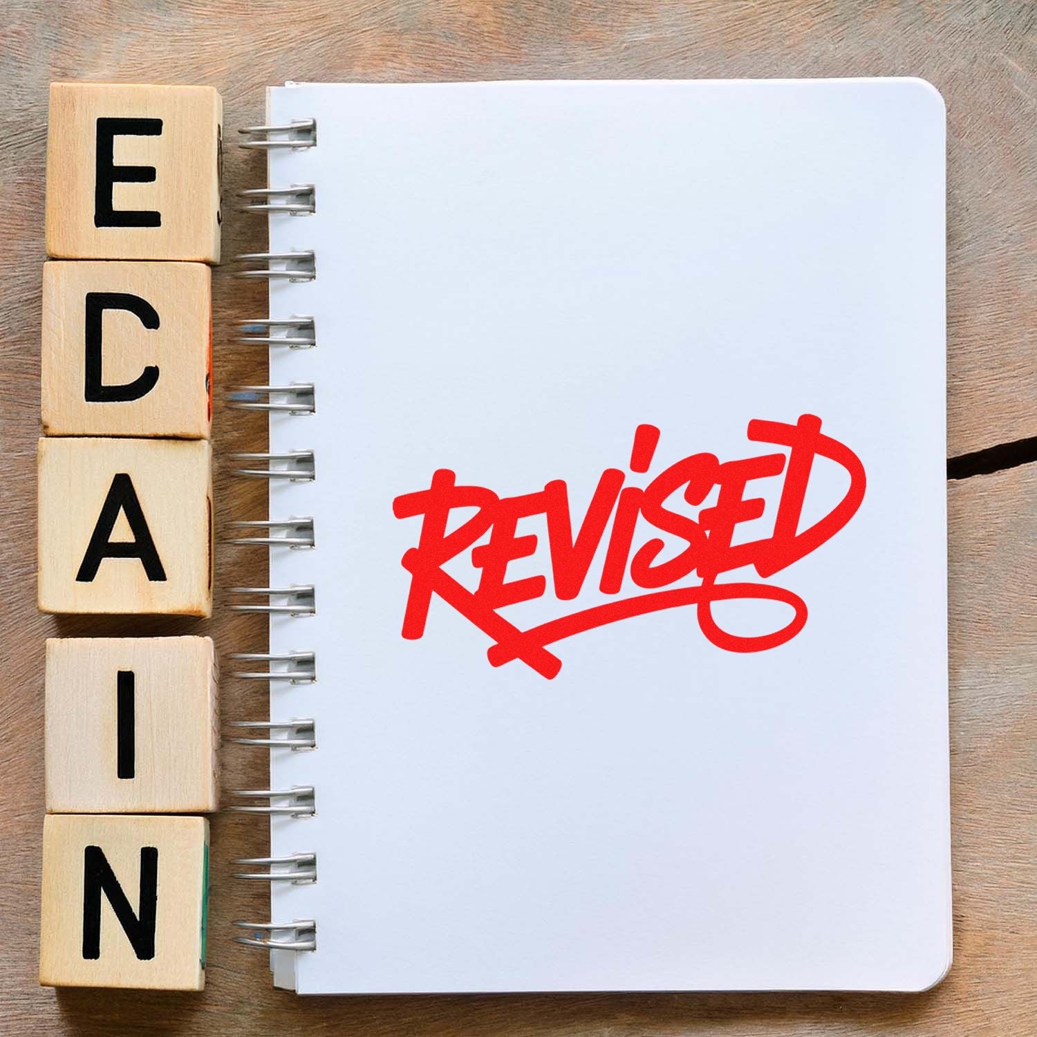 Wooden alphabet stamp blocks beside a white spiral notebook with a bold red revised seal imprint.