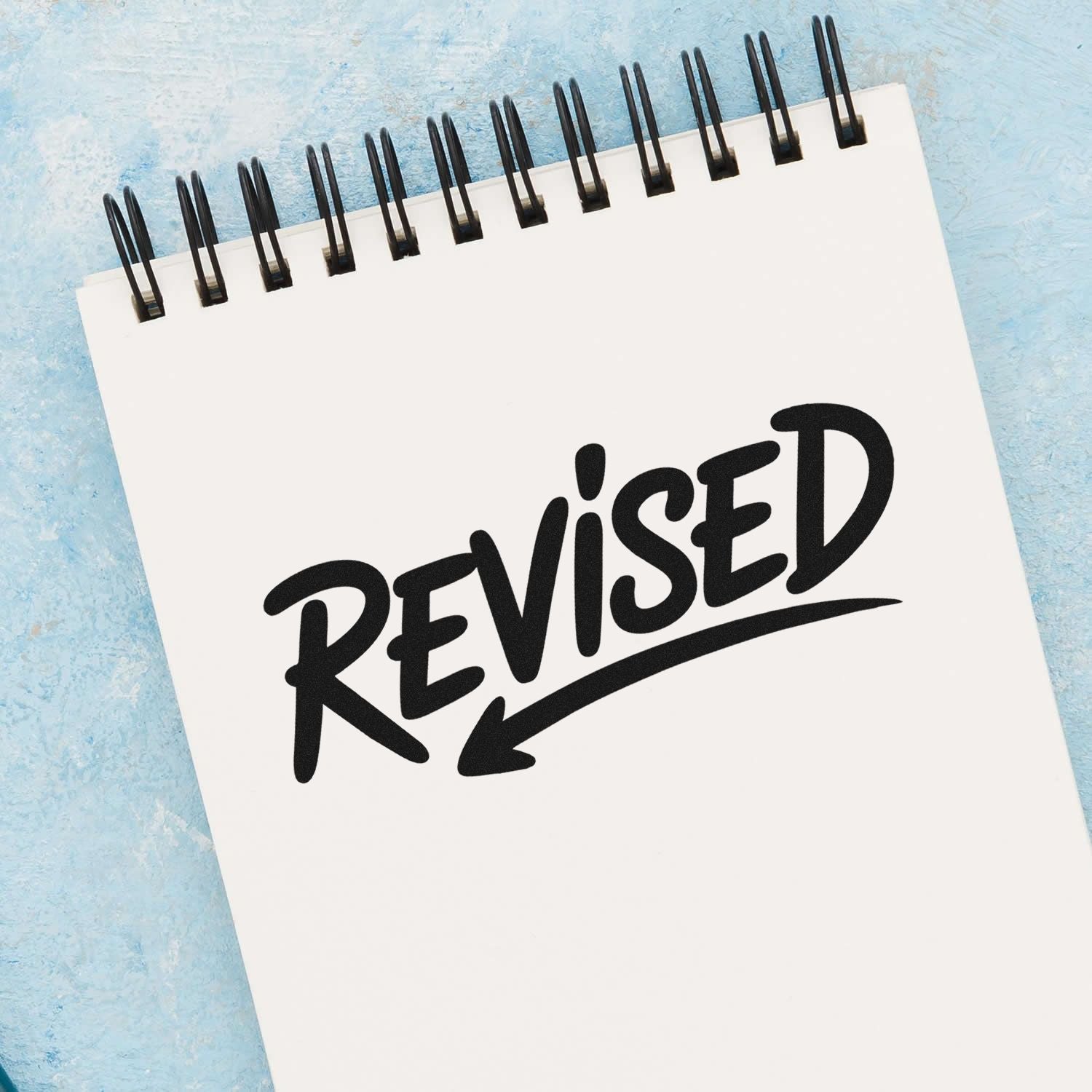 Black REVISED rubber stamp seal on a white spiral notepad, set against a light blue background.