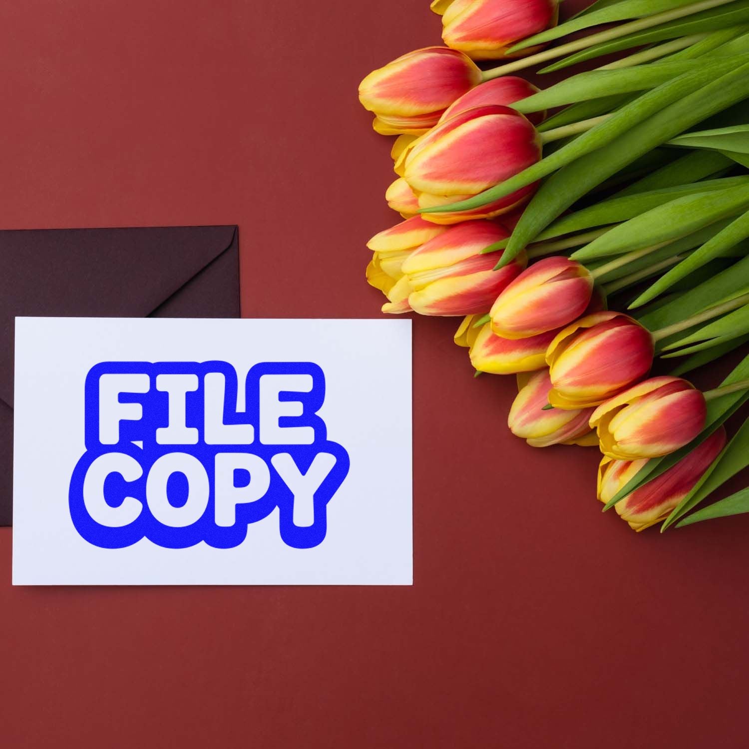 Blue FILE COPY stamp on white card beside black envelope and pink-yellow tulips on burgundy background.