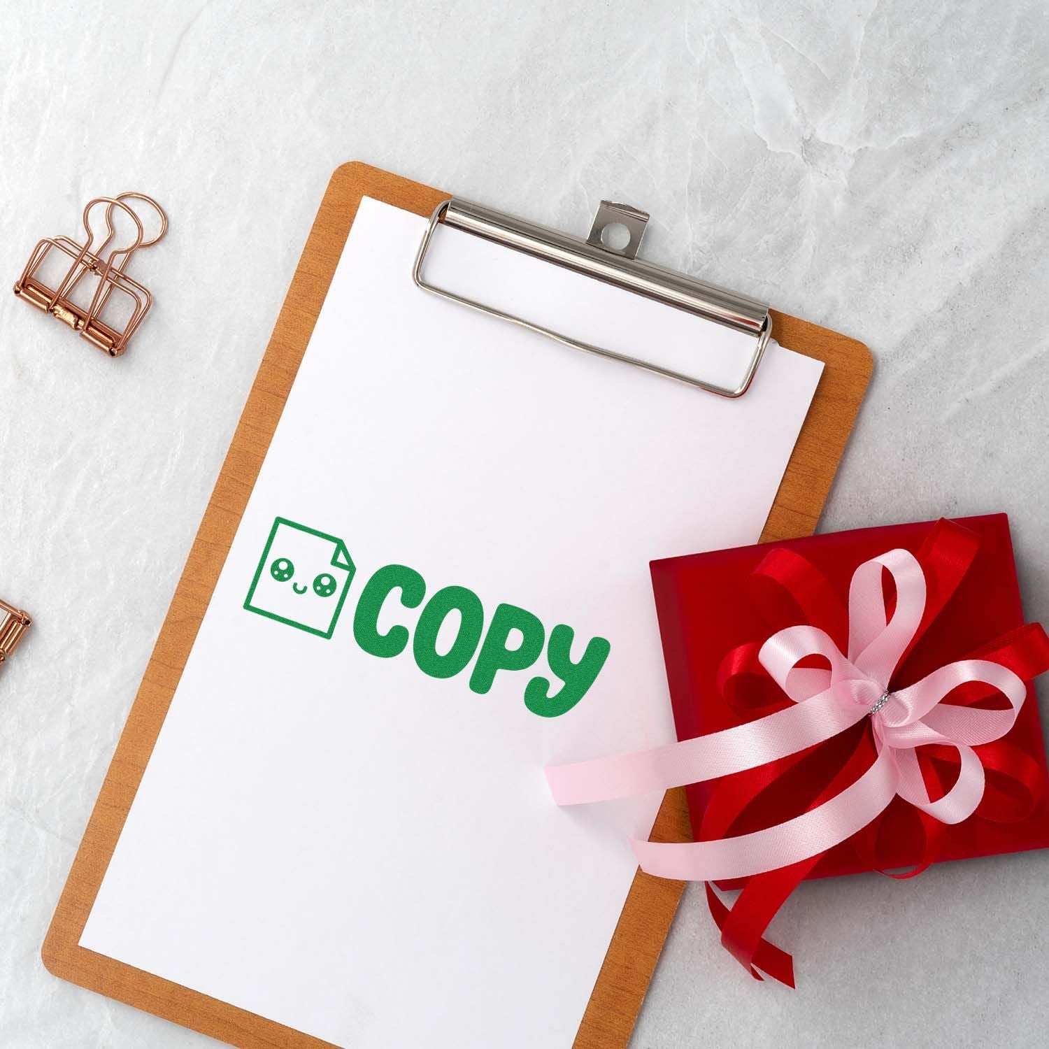 Clipboard displaying a green copy stamp, with a red gift seal and pink ribbon, on white marble background