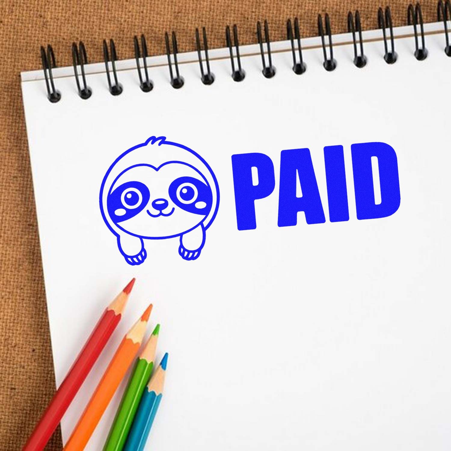 Blue PAID stamp and seal with cute sloth icon on a white spiral notebook, beside colorful pencils.
