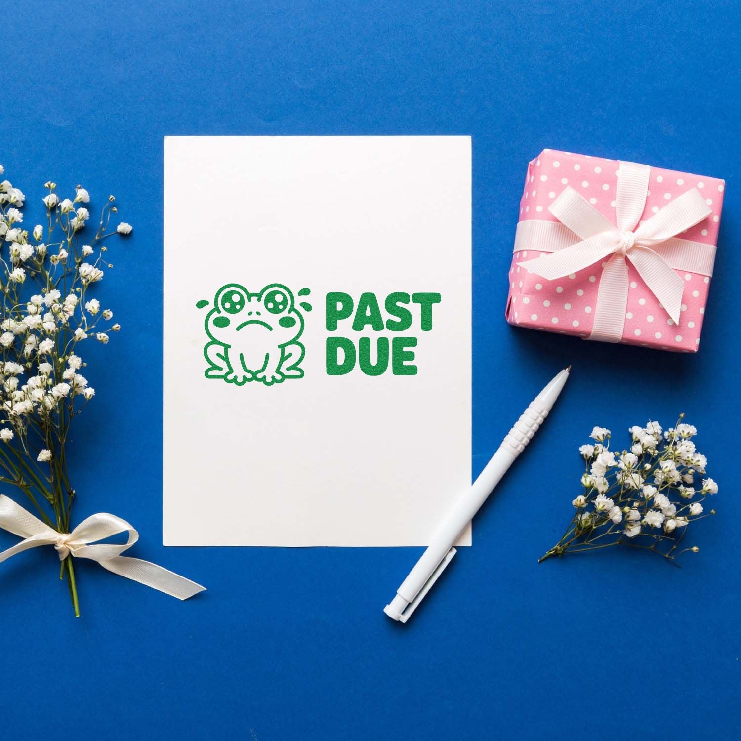 Green PAST DUE frog stamp on white card, with pink polka-dot gift box, white pen, and white flowers on blue.