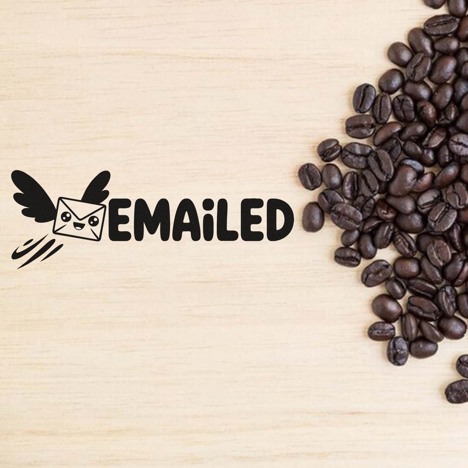 Black Emailed rubber stamp seal with winged envelope icon on light wood, beside dark coffee beans