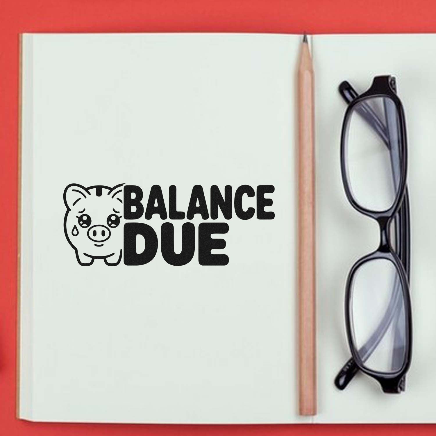Black BALANCE DUE stamp and seal with pig icon on white notebook, beside pencil and eyeglasses, on red background