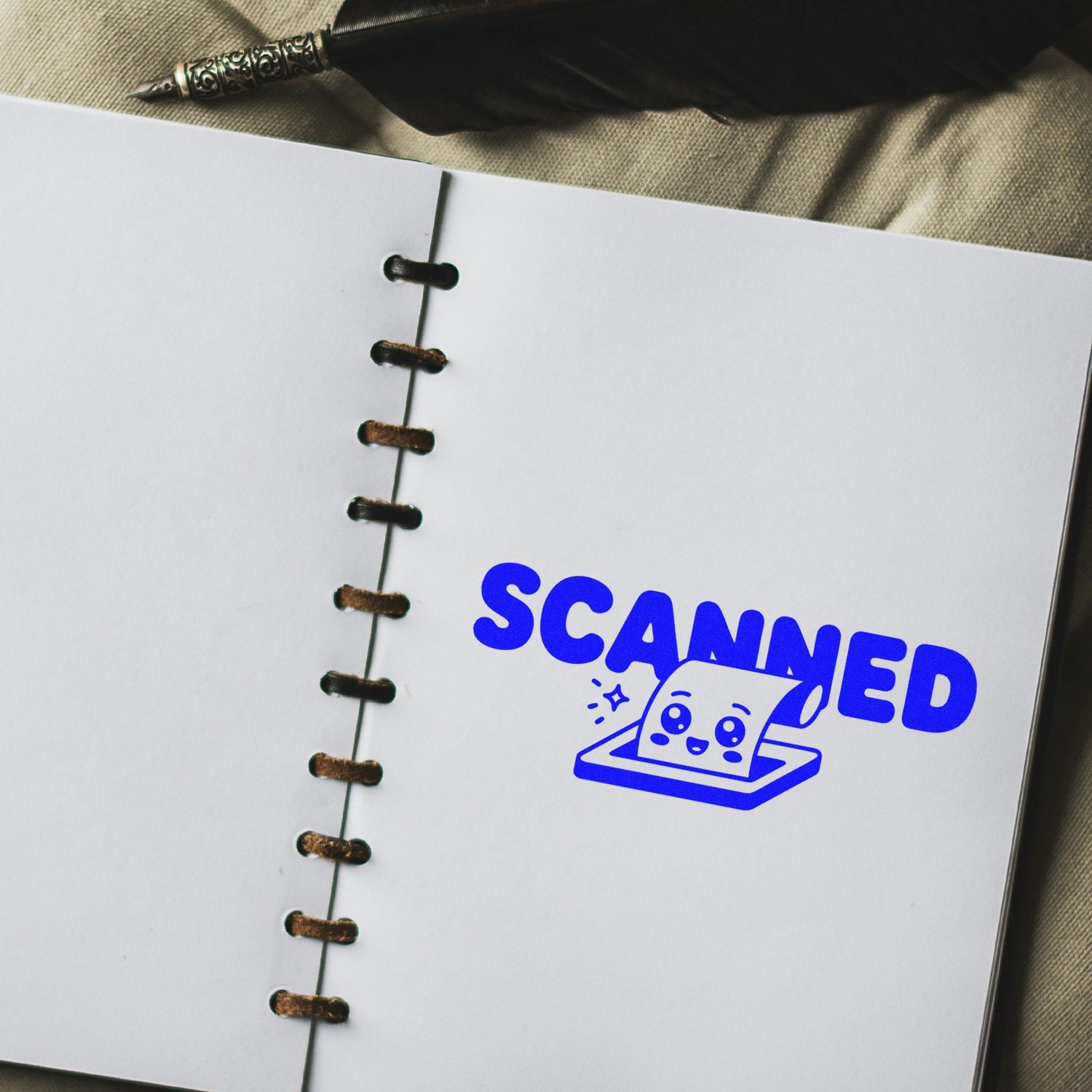 White spiral notebook with bold blue SCANNED stamp and cute scanner seal on beige fabric, beside a pen and quill.